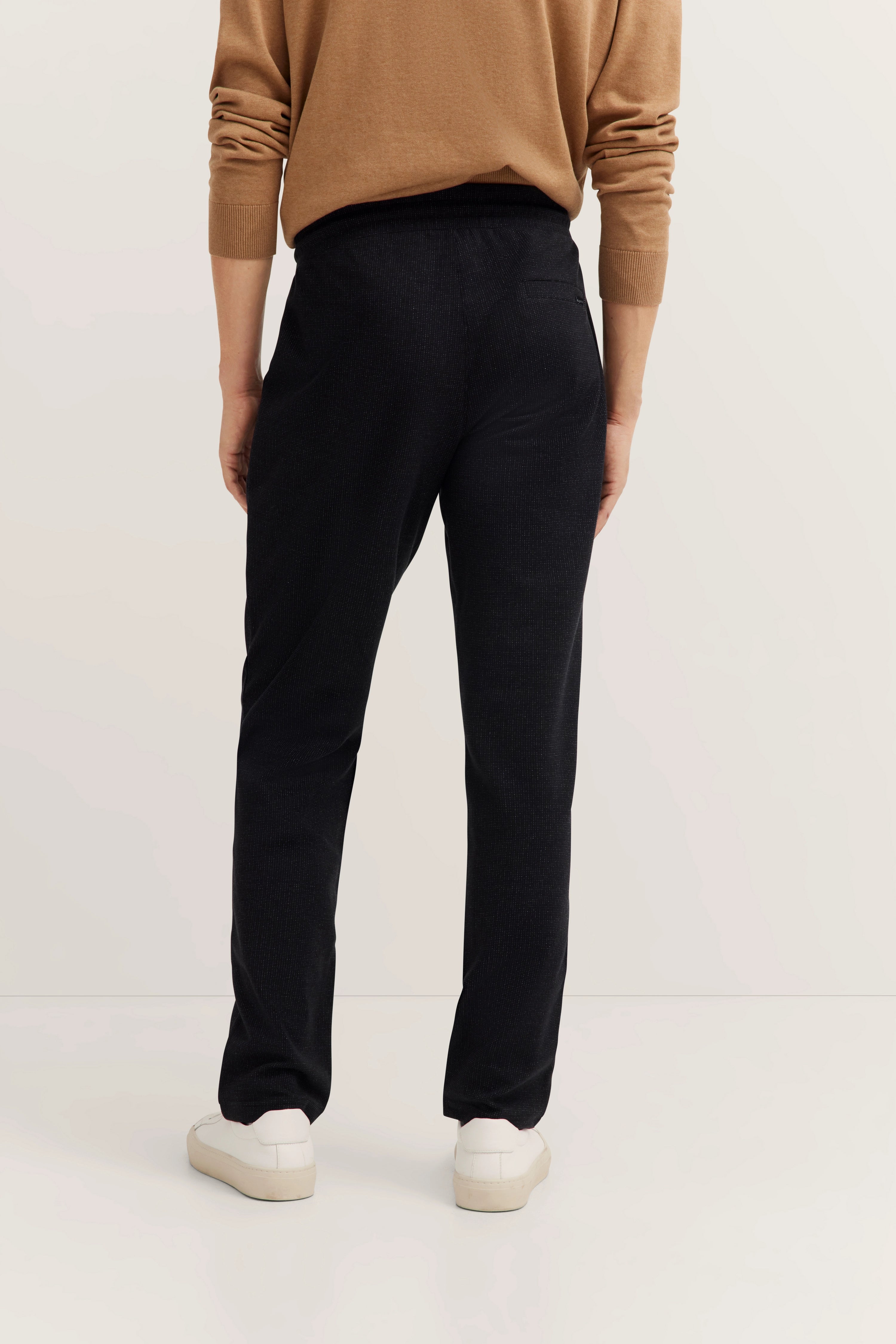 Bequemes Modelbild mit camel Pullover, schwarzer Hose und weißen Sneakern von bugatti, Rückansicht.