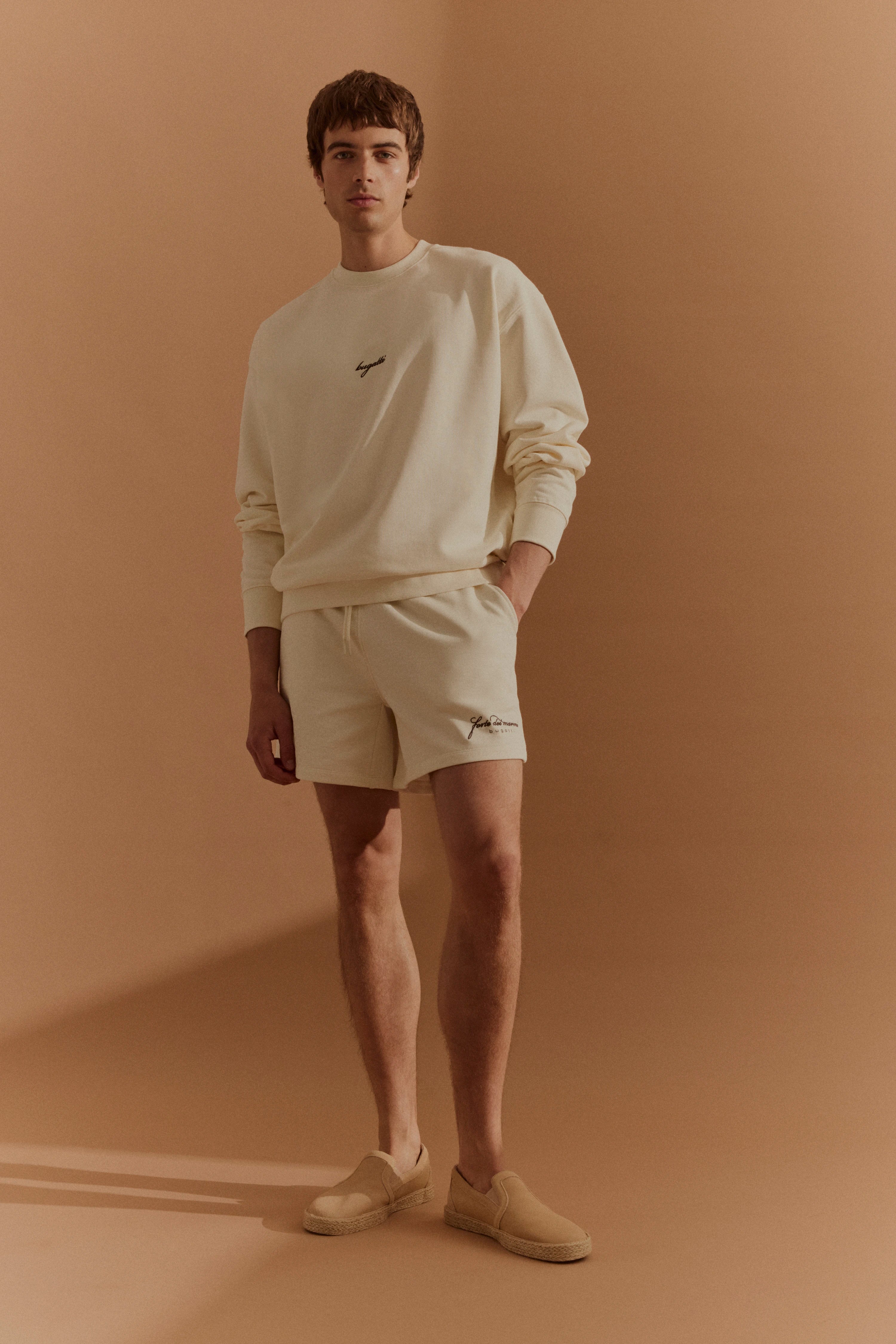 Model in cremefarbenem bugatti Sweatshirt und Shorts, beige Slip-Ons, entspannte Pose.