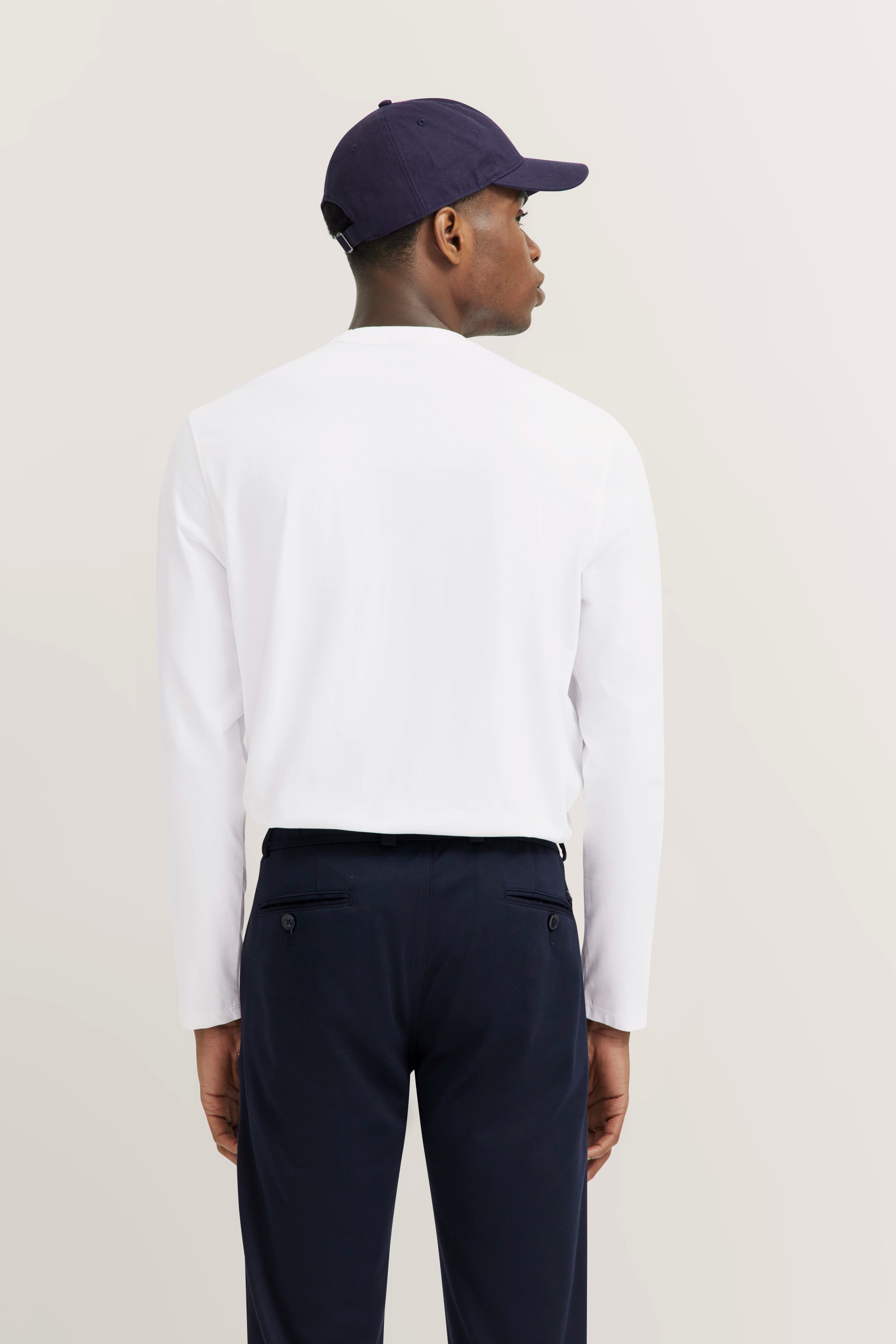 Model from behind in a white long-sleeve shirt and dark blue pants, wearing a dark blue cap.
