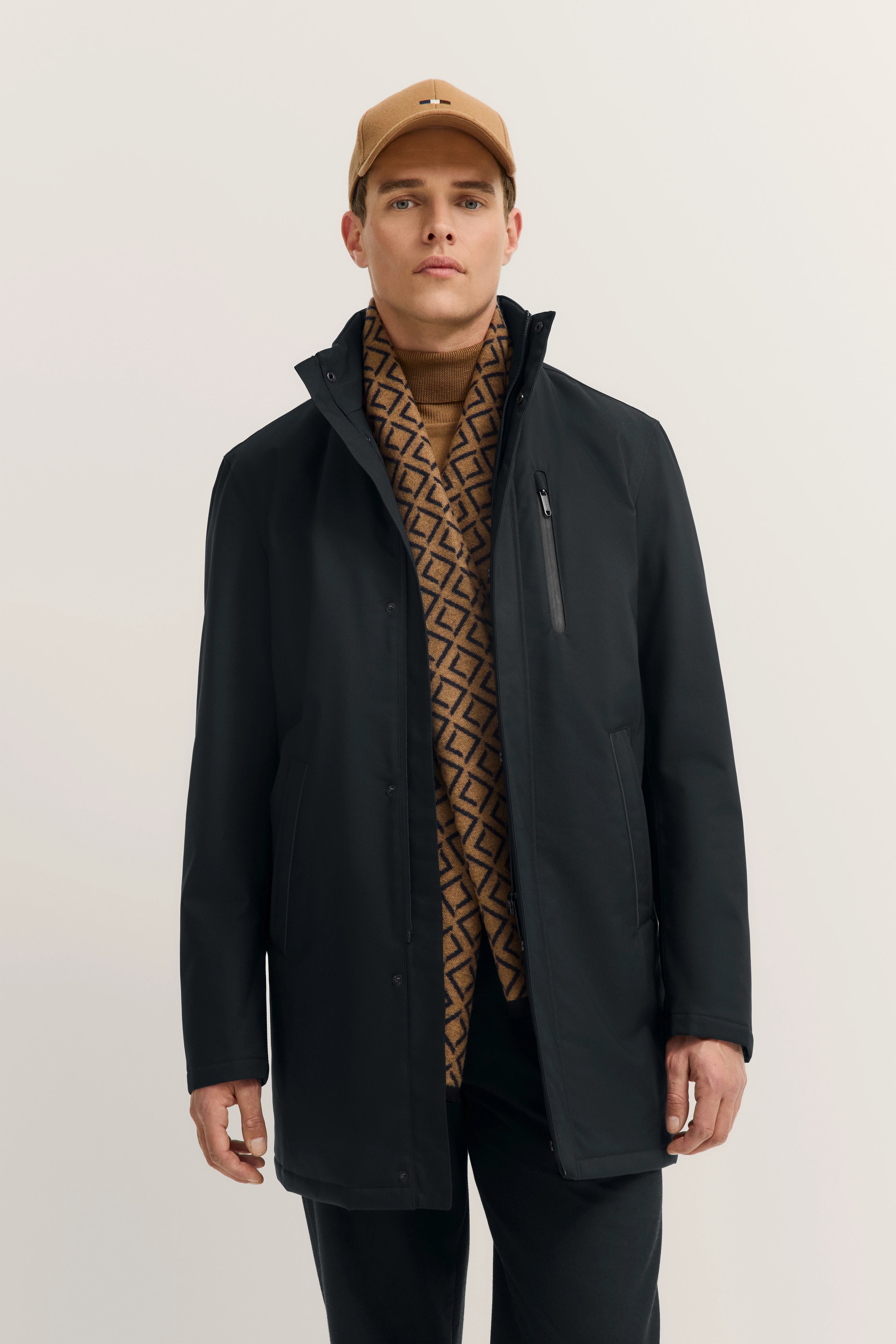 Model in schwarzem Mantel, braunem Schal mit Muster und beiger Kappe von bugatti, frontal stehend.