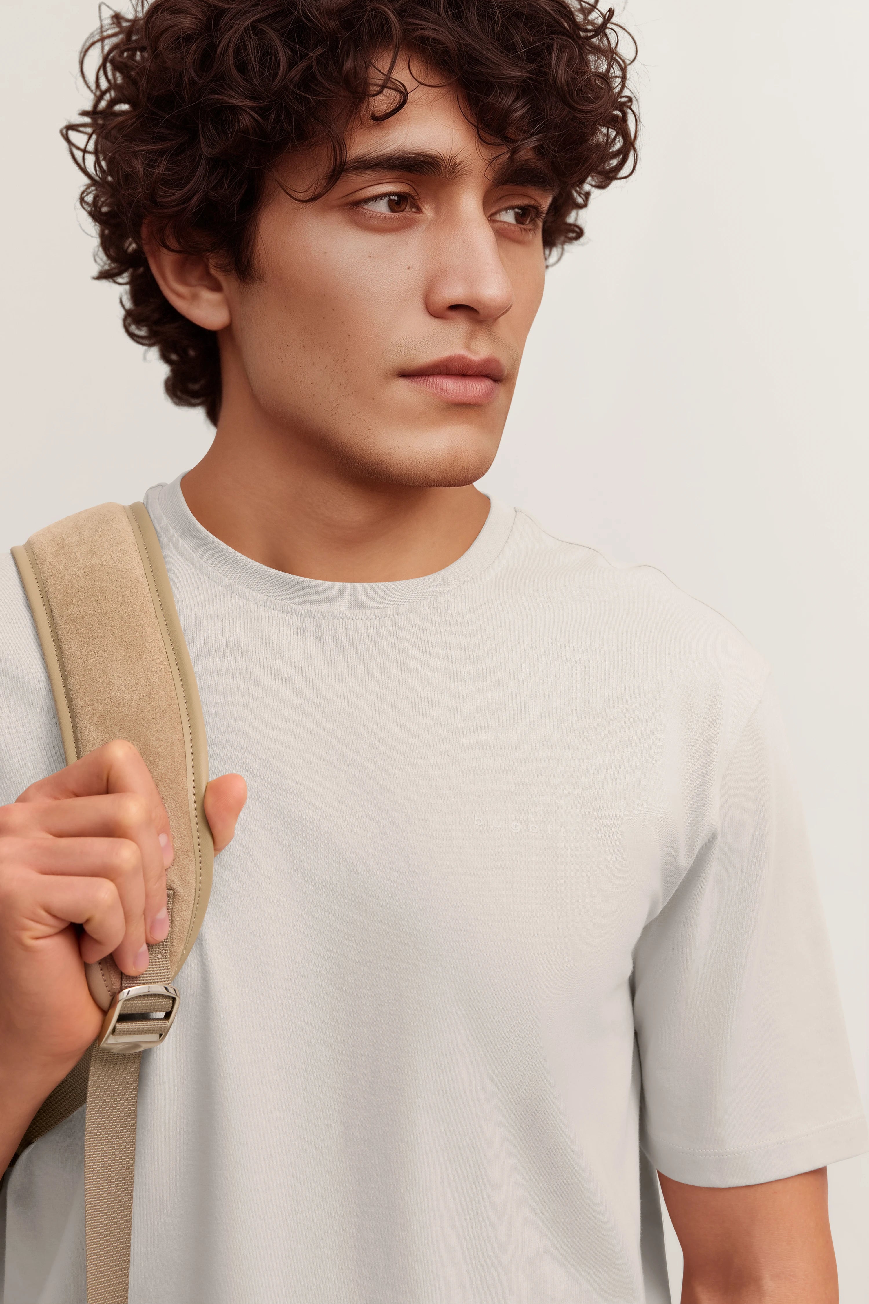 bugatti Modelbild, weißes T-Shirt, beigefarbener Rucksack, entspannte Pose, stilvoll lässig.