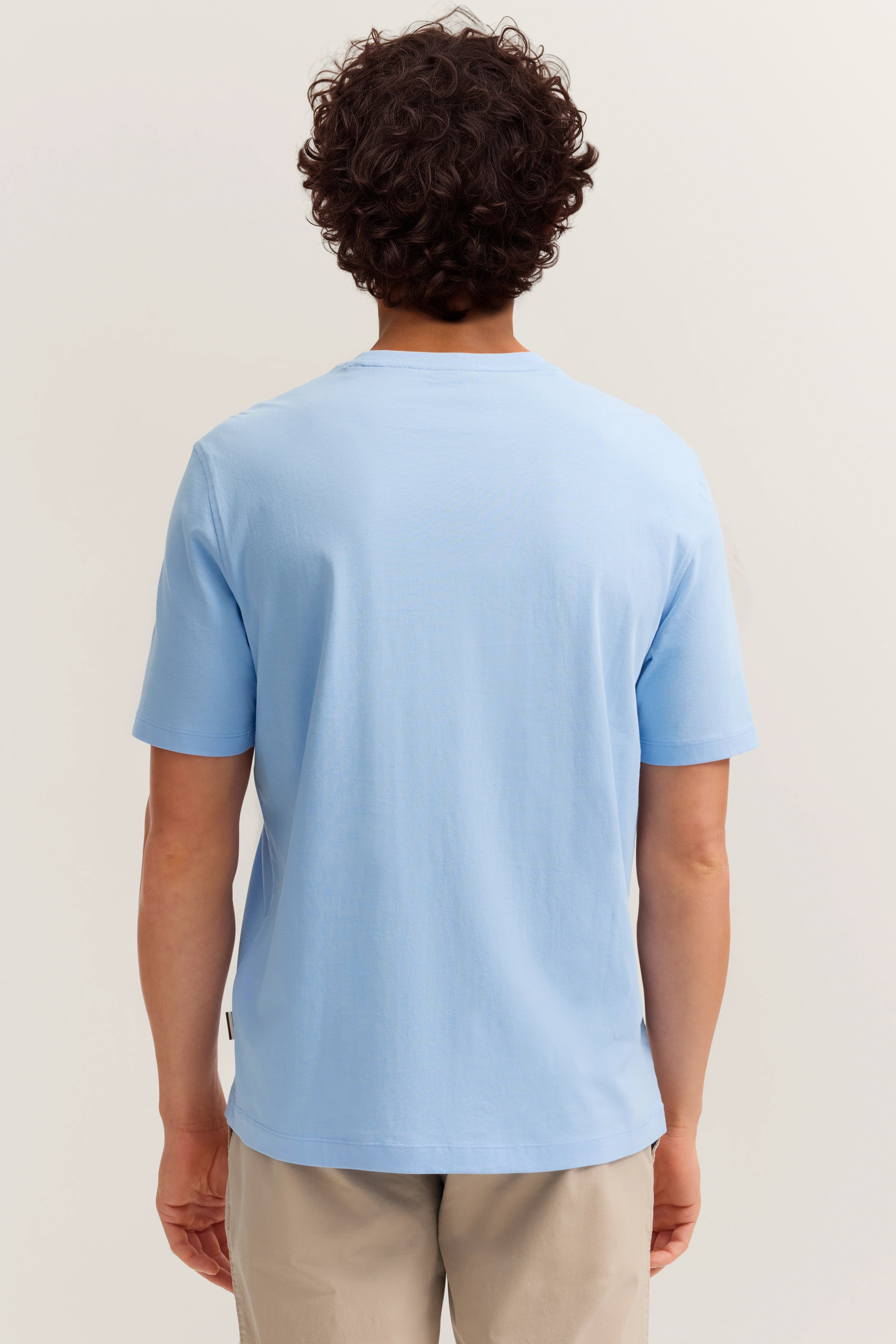 Rückenansicht eines legeren Models in hellblauem bugatti T-Shirt und beigen Hosen.