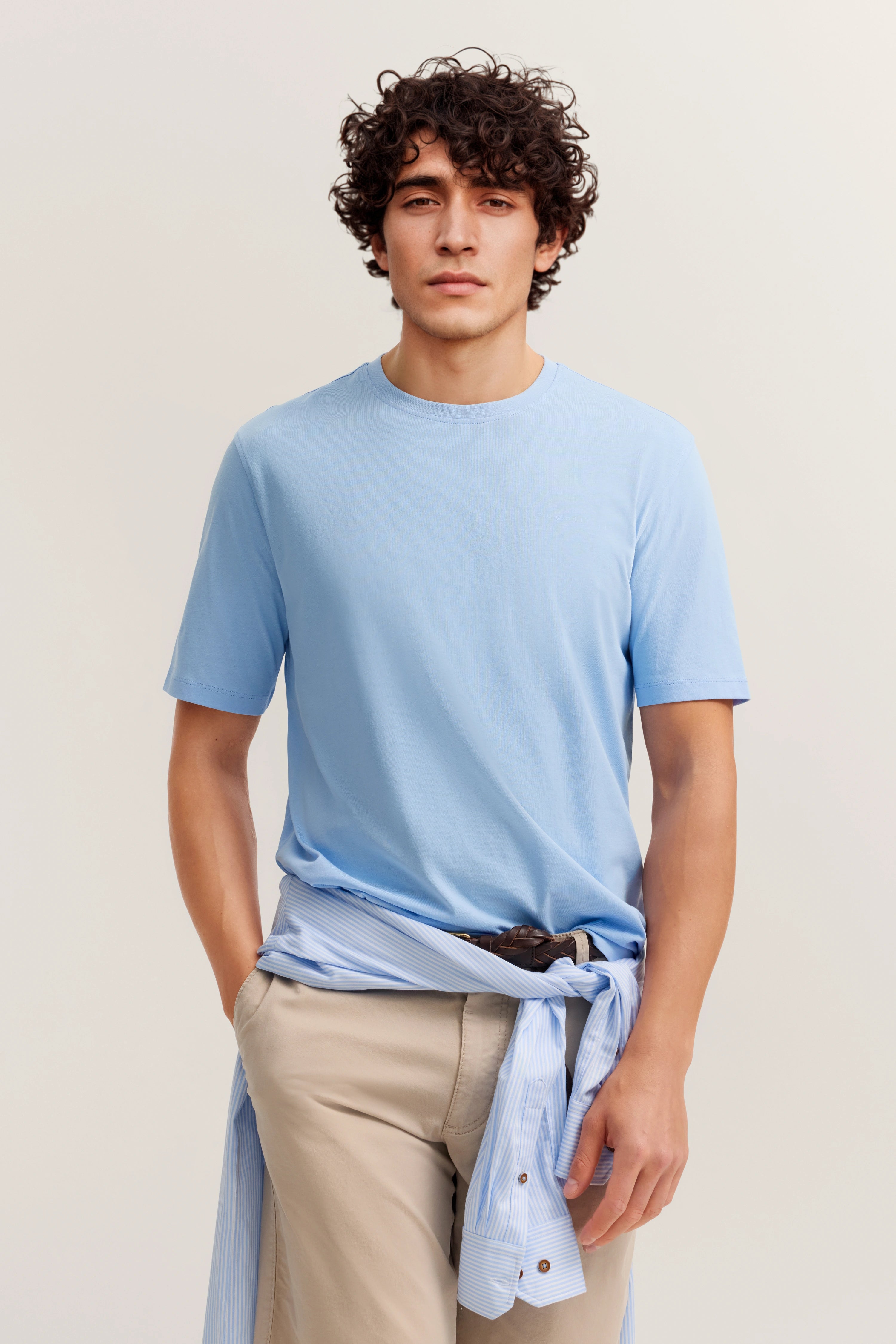 Blaues T-Shirt, beige Hose, gestreiftes Hemd um die Taille, legeres Modelbild, entspannte Pose.