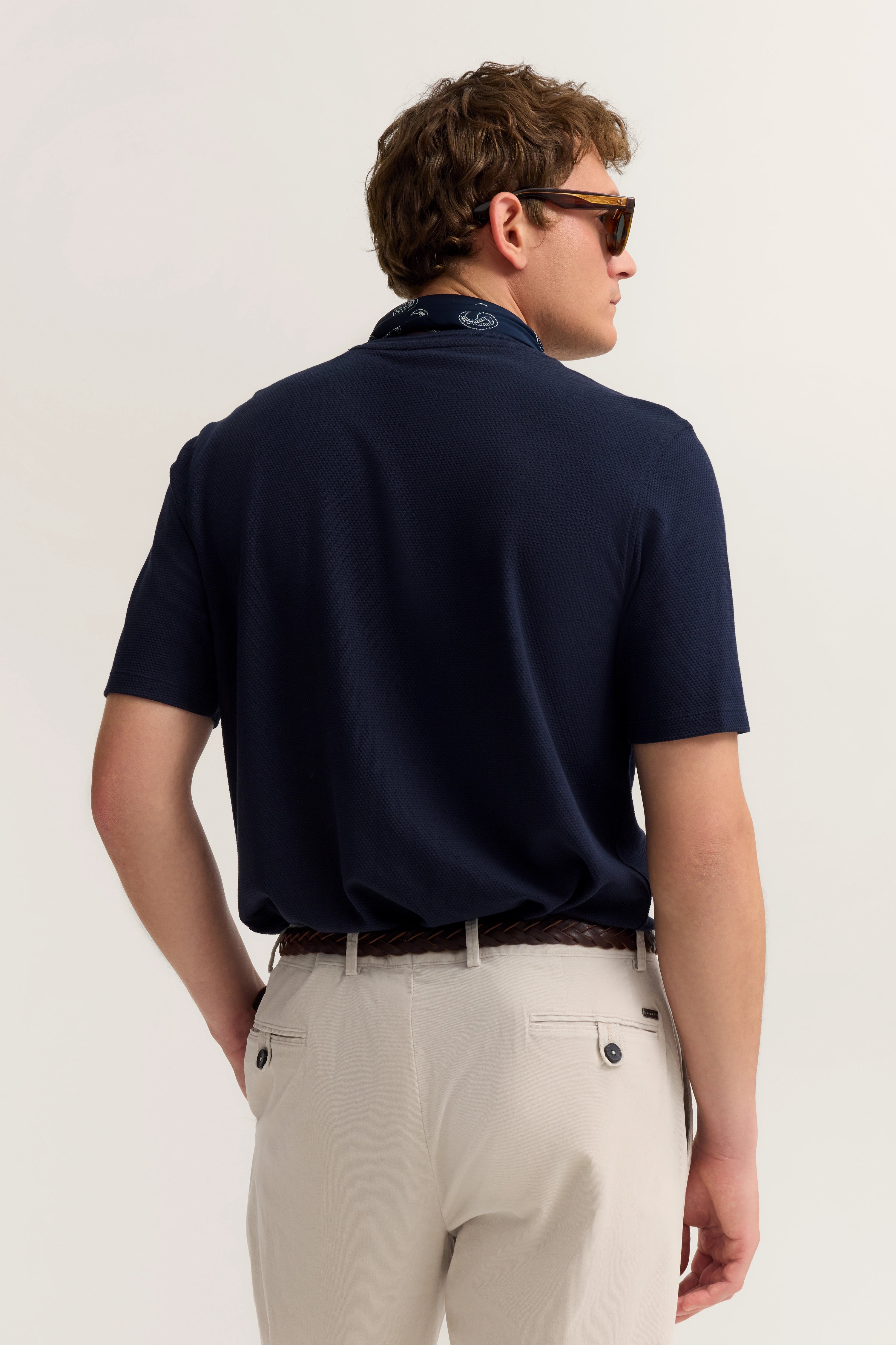 bugatti Modelbild, navy Polo, beige Chinos, brauner Gürtel, seitliches Posing, Sonnenbrille, Rückansicht.