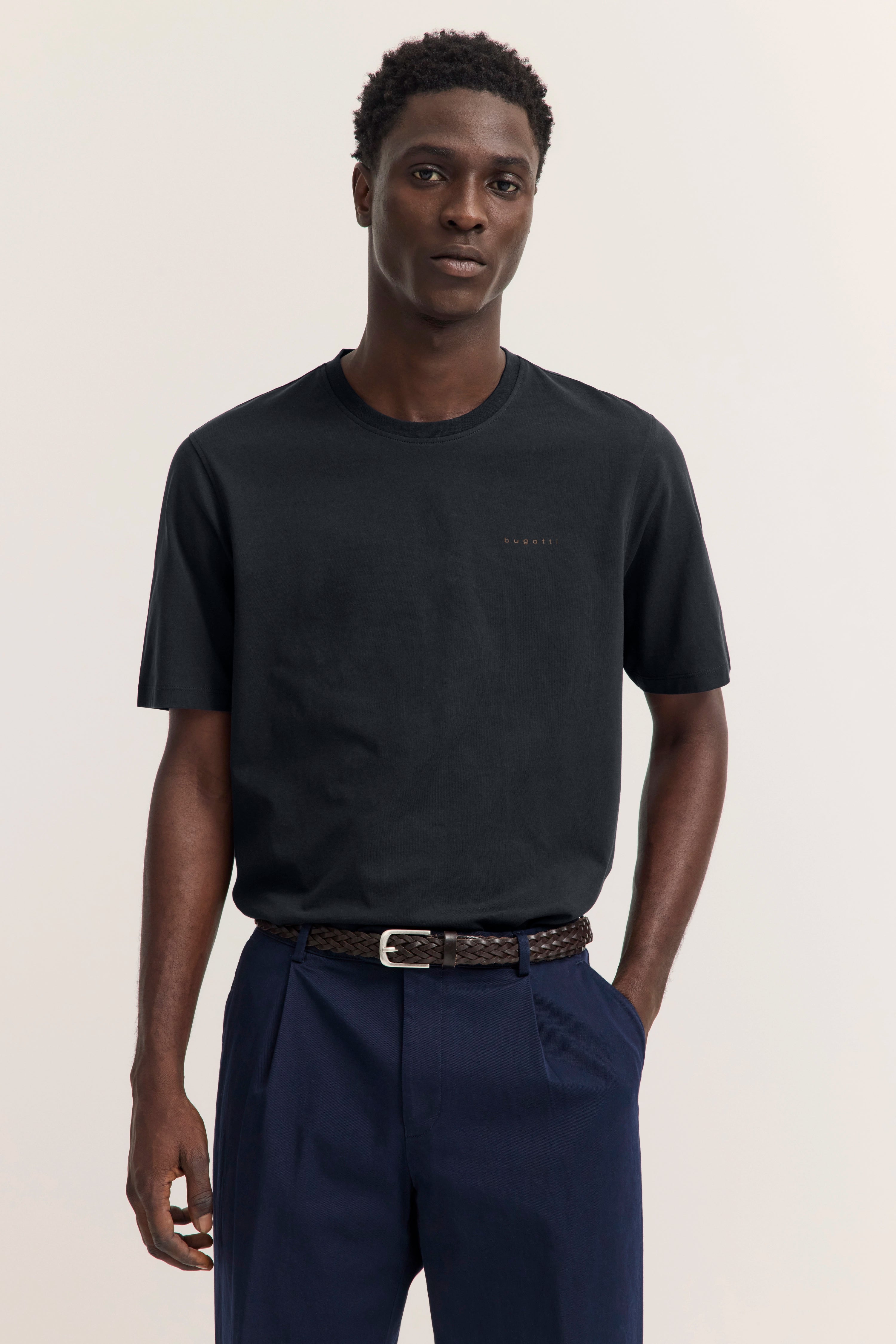 bugatti schwarzes T-Shirt und blaue Hose, Model steht entspannt, Hände in Taschen.