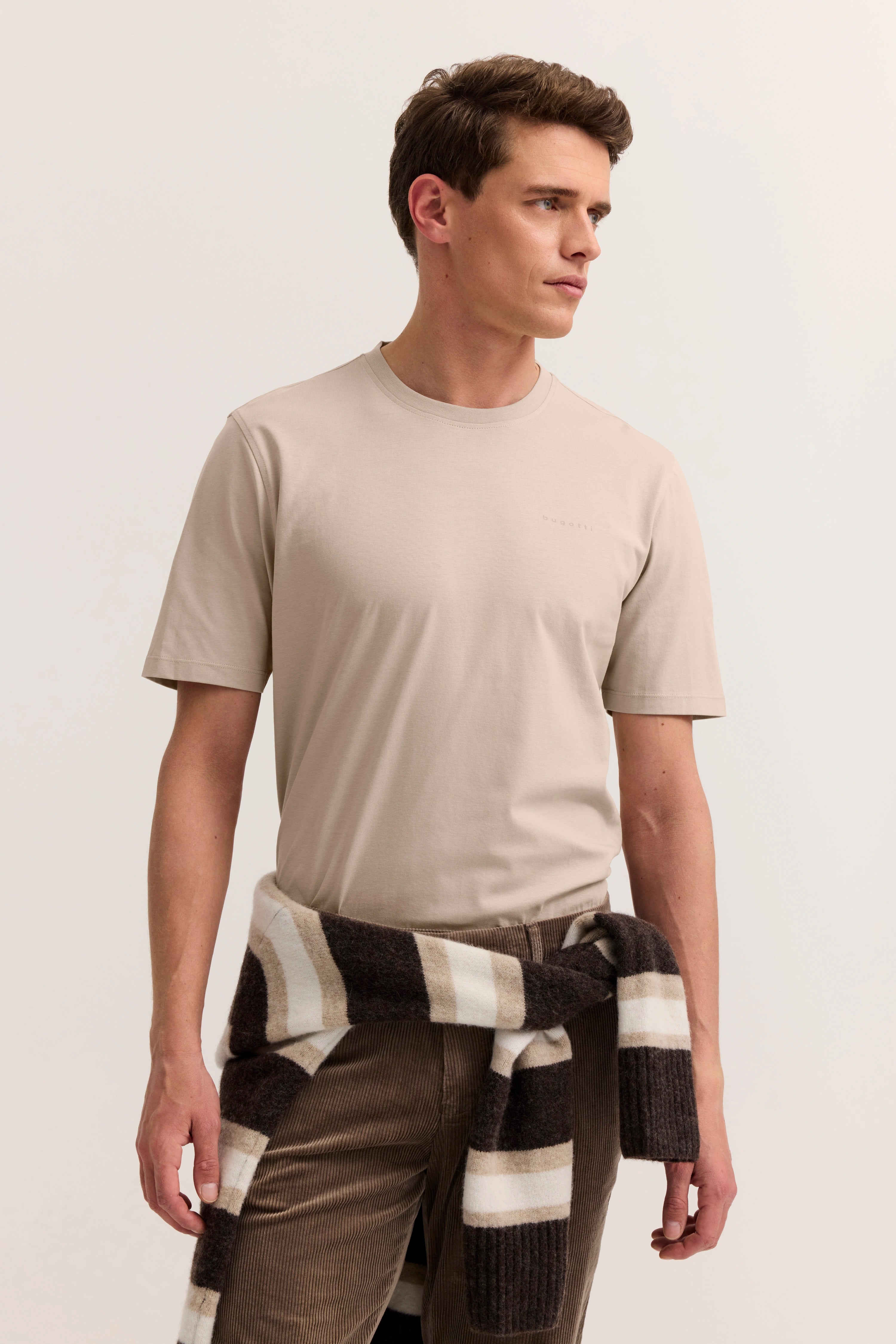 Beiges bugatti T-Shirt, braune Cordhose, gestreifter Pullover um die Taille. Model seitlich blickend.