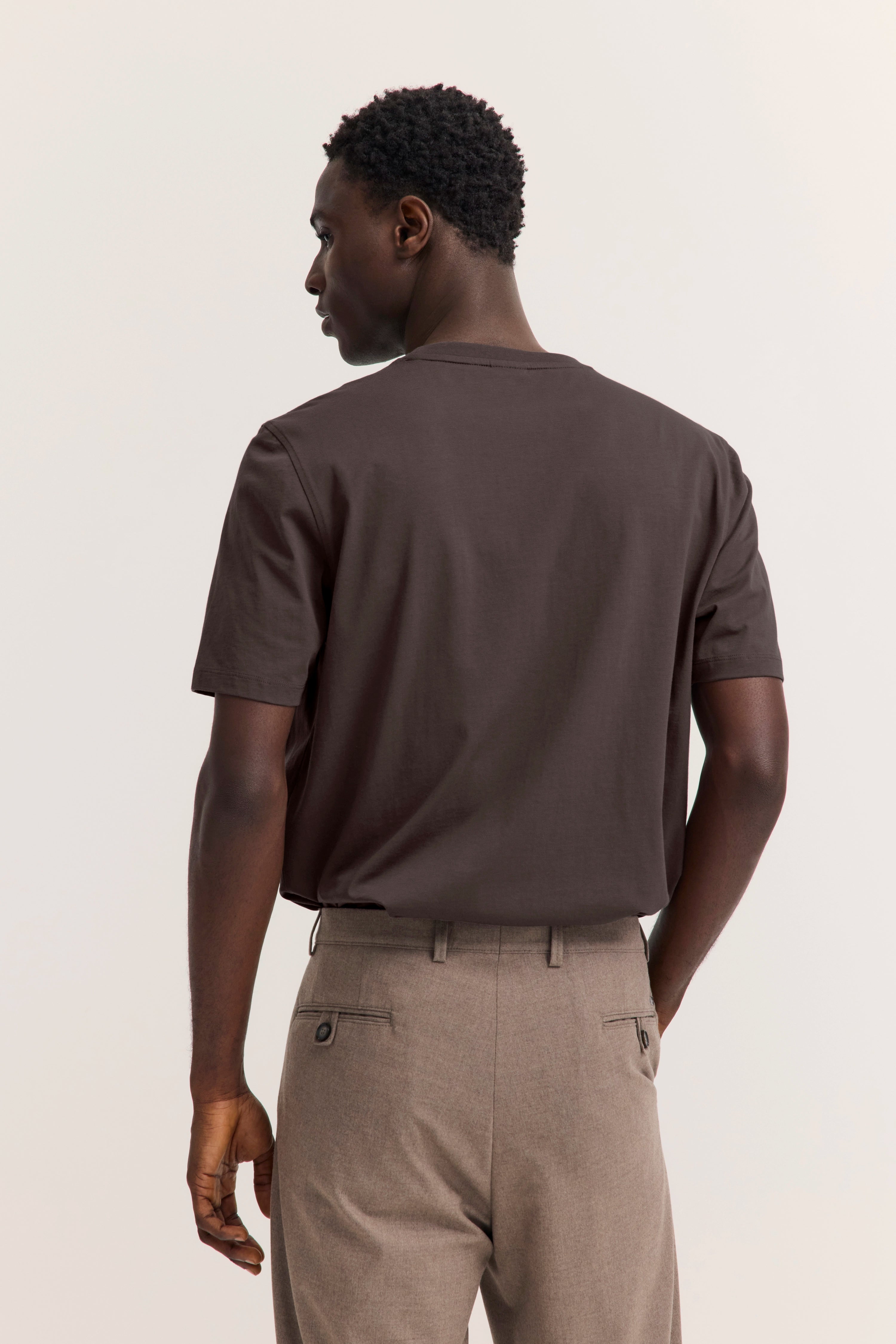 Herrenmodell in braunem T-Shirt und beiger Hose von hinten, lässiger Stand.