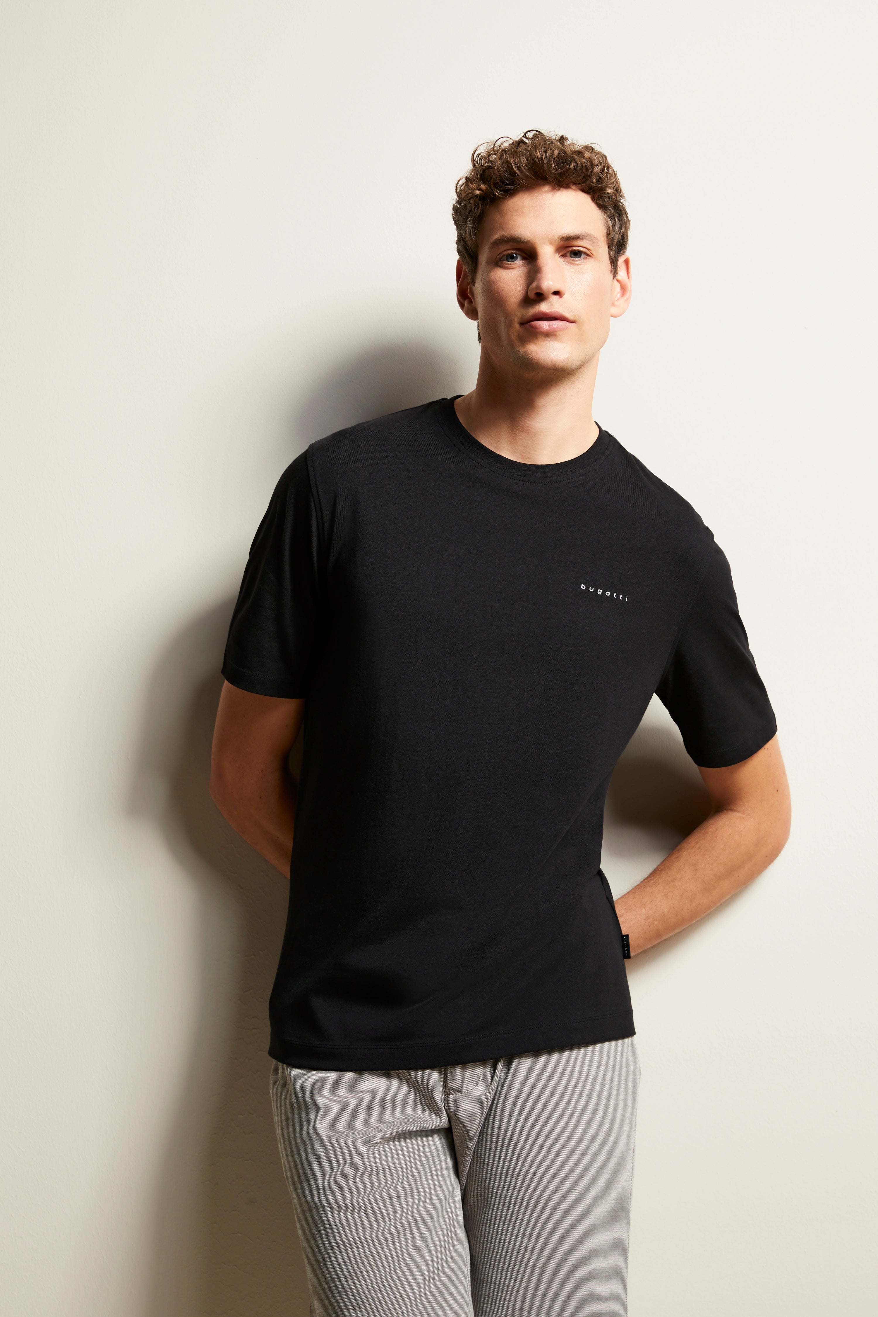 Black bugatti T-shirt, gray pants, model pose, casual with arms behind back.