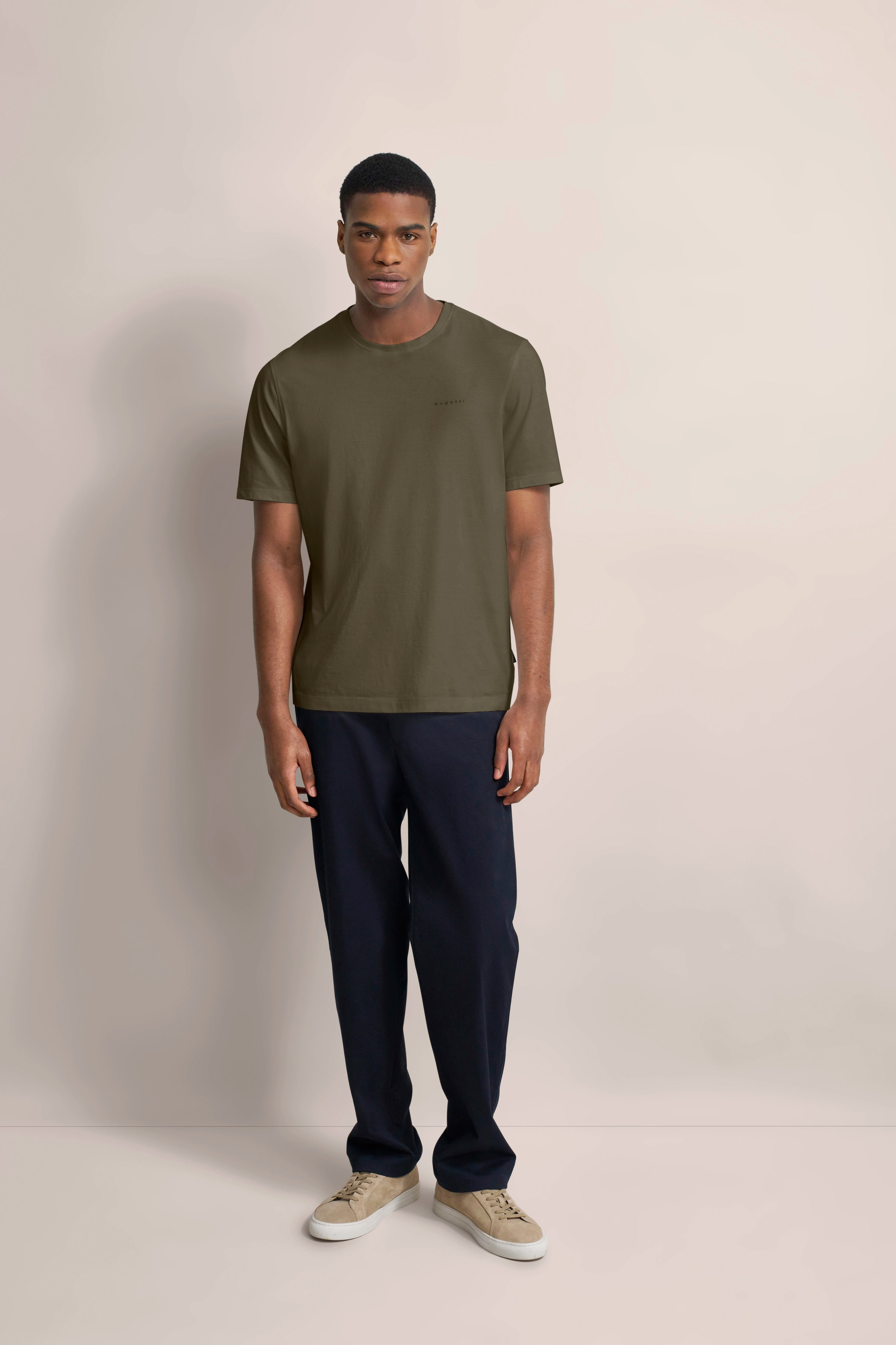 Model in olivgrünem bugatti T-Shirt, dunkelblauer Hose, beige Sneaker, entspanntes Posing.