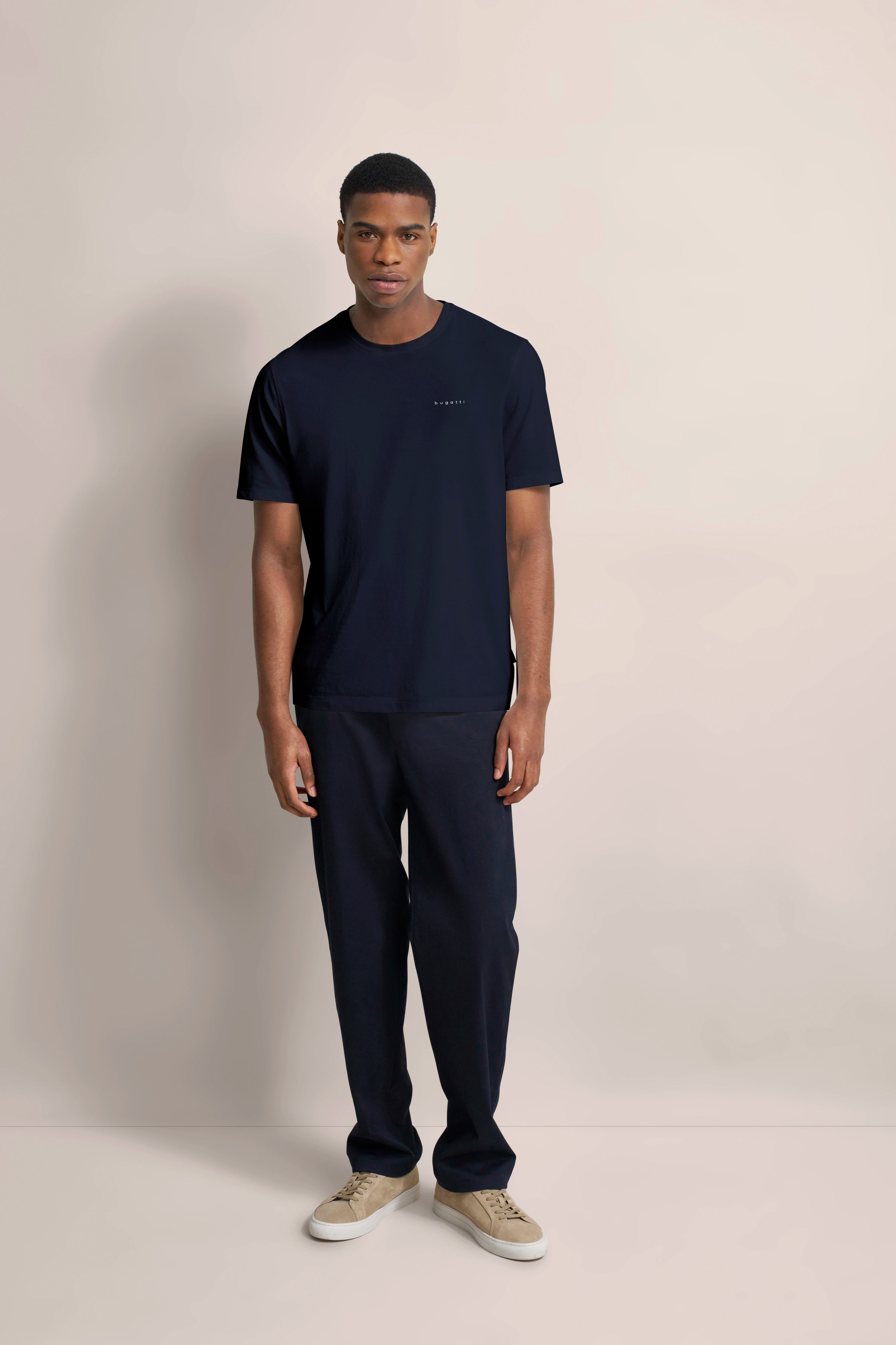 bugatti Outfit: schwarzes T-Shirt, schwarze Hose, beiges Schuhwerk. Modelbild, entspanntes Stehen.