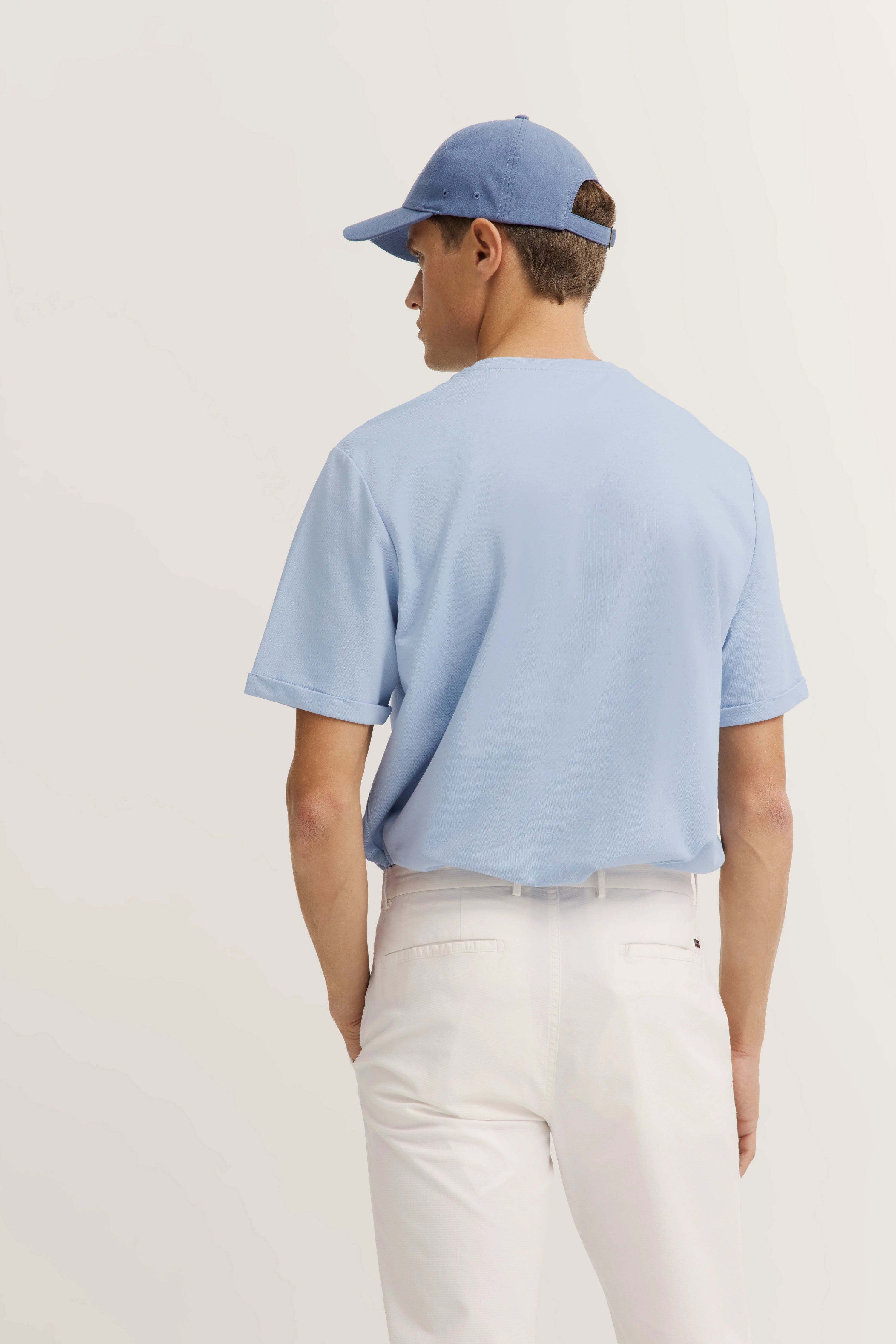 Model in hellblauem Shirt und weißer Hose, trägt passende Kappe, Rückenansicht.