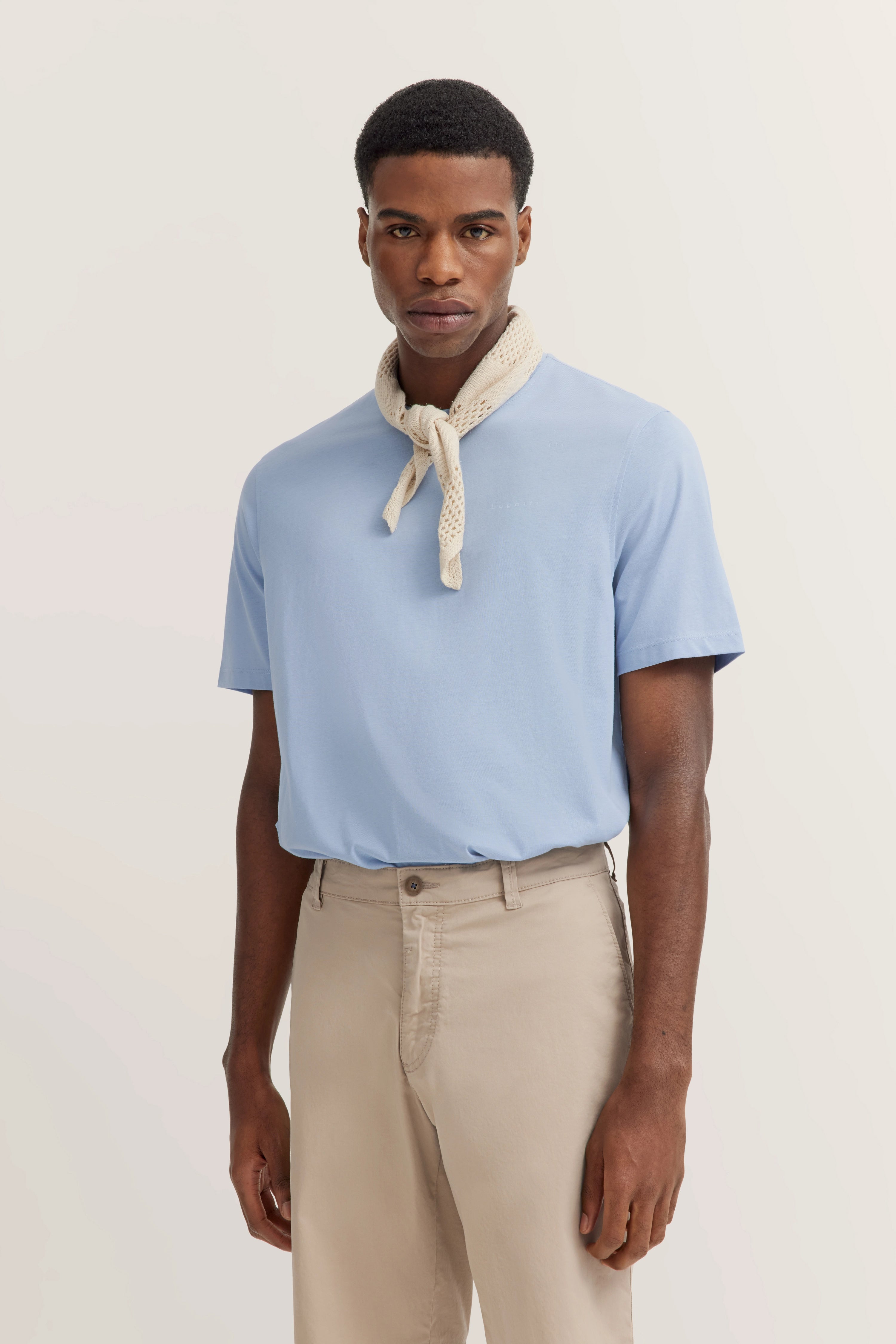 Modell in hellblauem Shirt, beigefarbene Hose, beiges Halstuch. Stehend, entspannter Ausdruck.