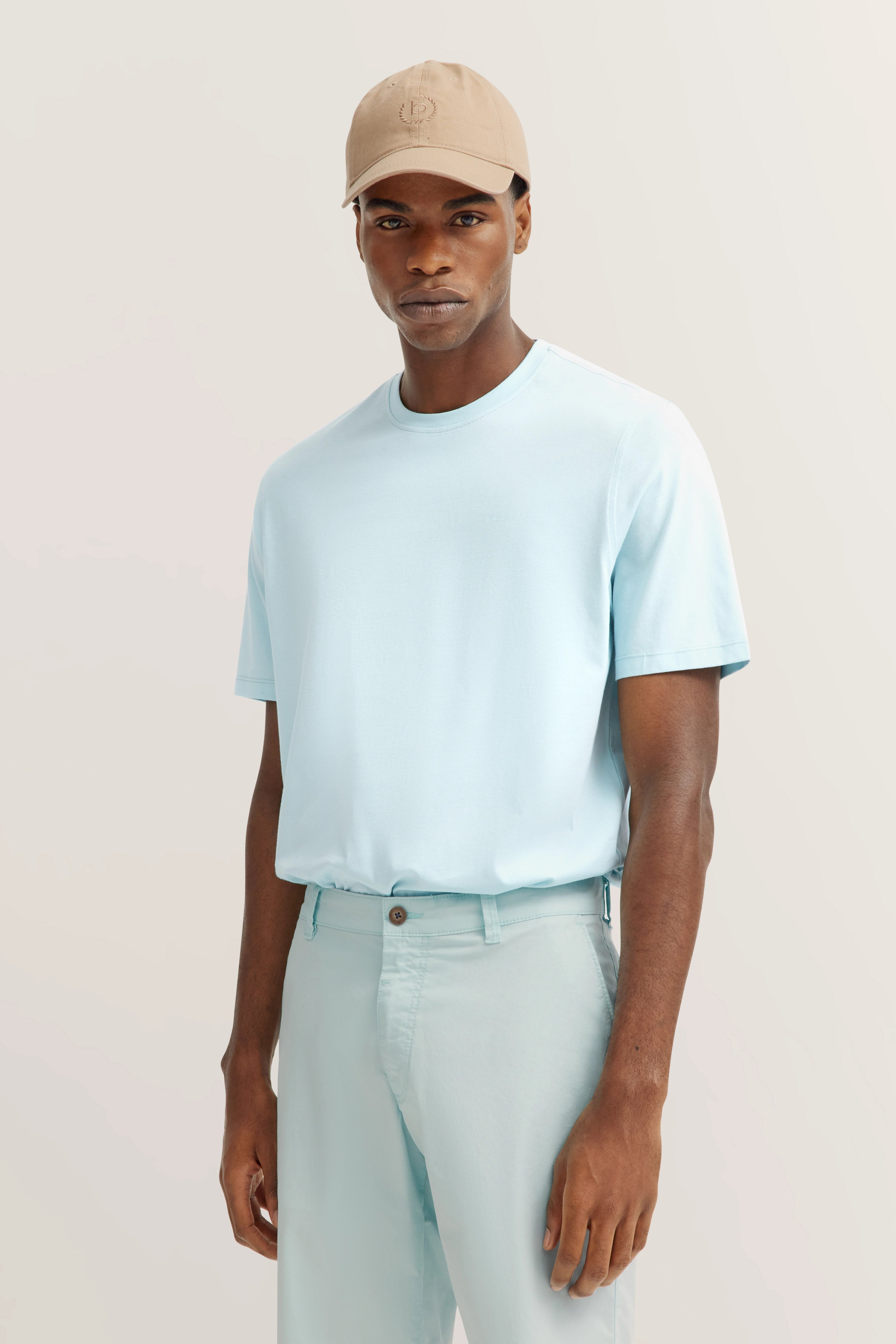 Model in hellblauem bugatti T-Shirt und Hose, beige Kappe, entspanntes Posing, Halbfiguraufnahme.