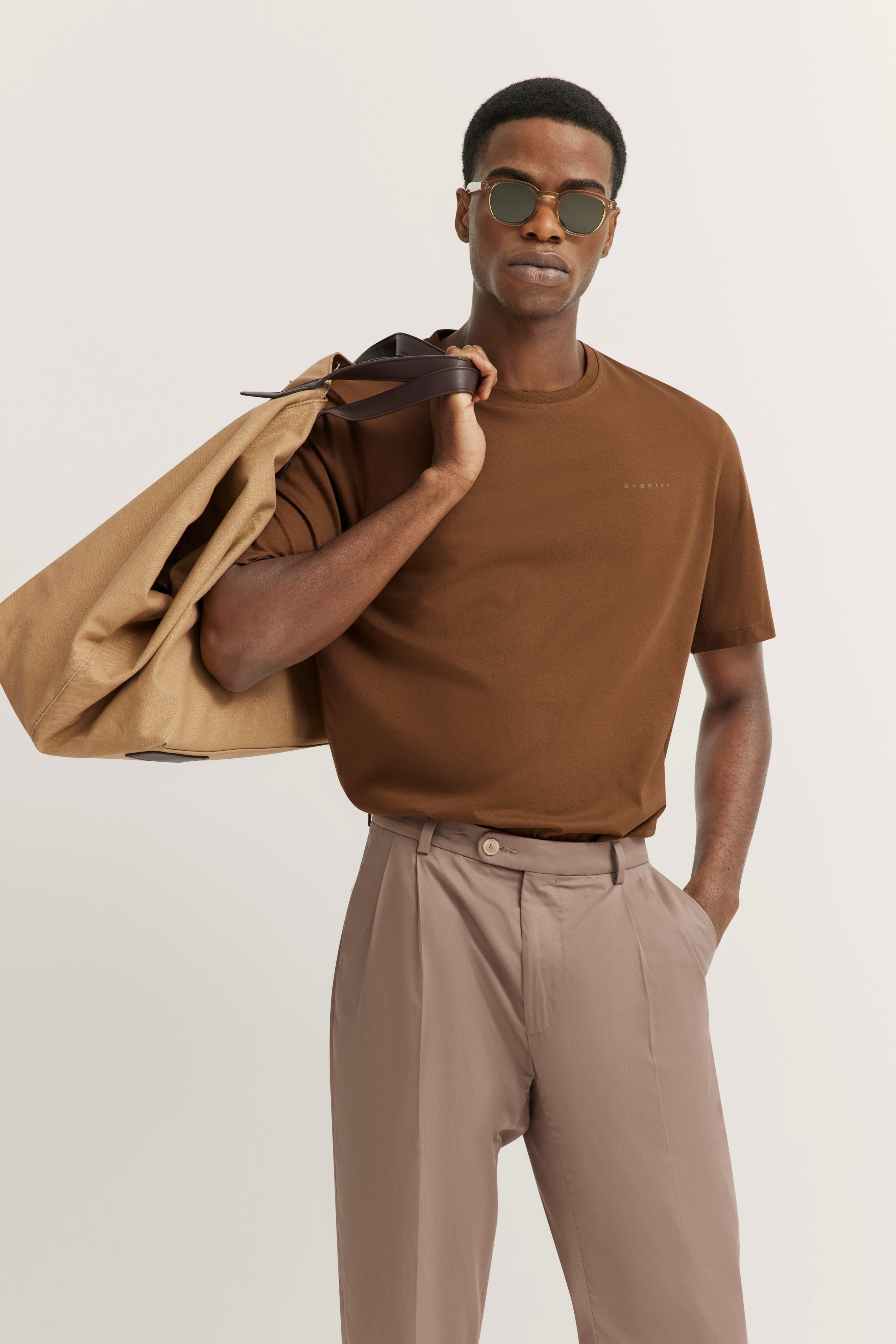 Model in brown bugatti T-shirt, beige pants, sunglasses, wearing beige coat over shoulder.