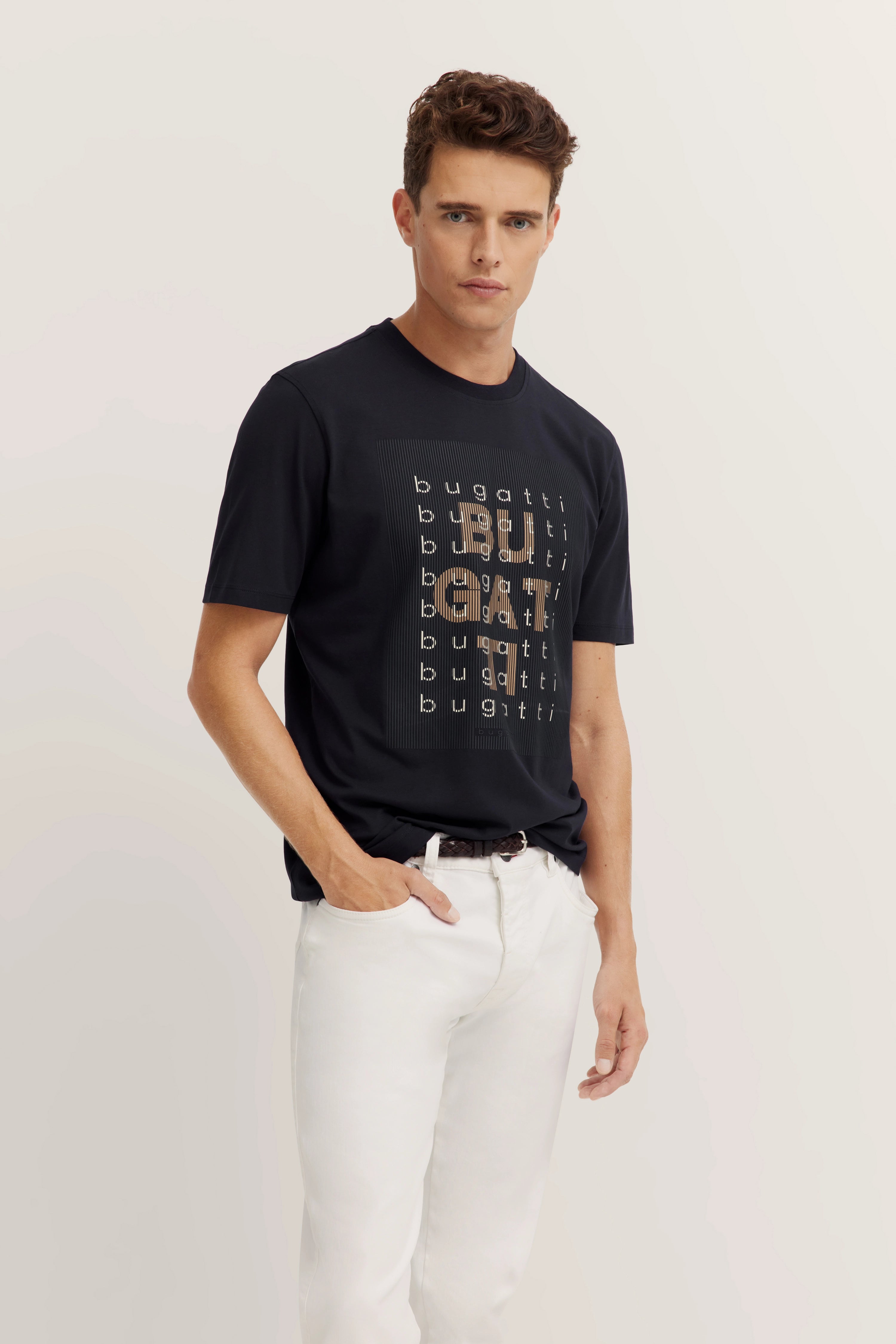 Model in schwarzem bugatti T-Shirt mit Logo, weiße Hose, lässige Pose, Hand in Hosentasche.