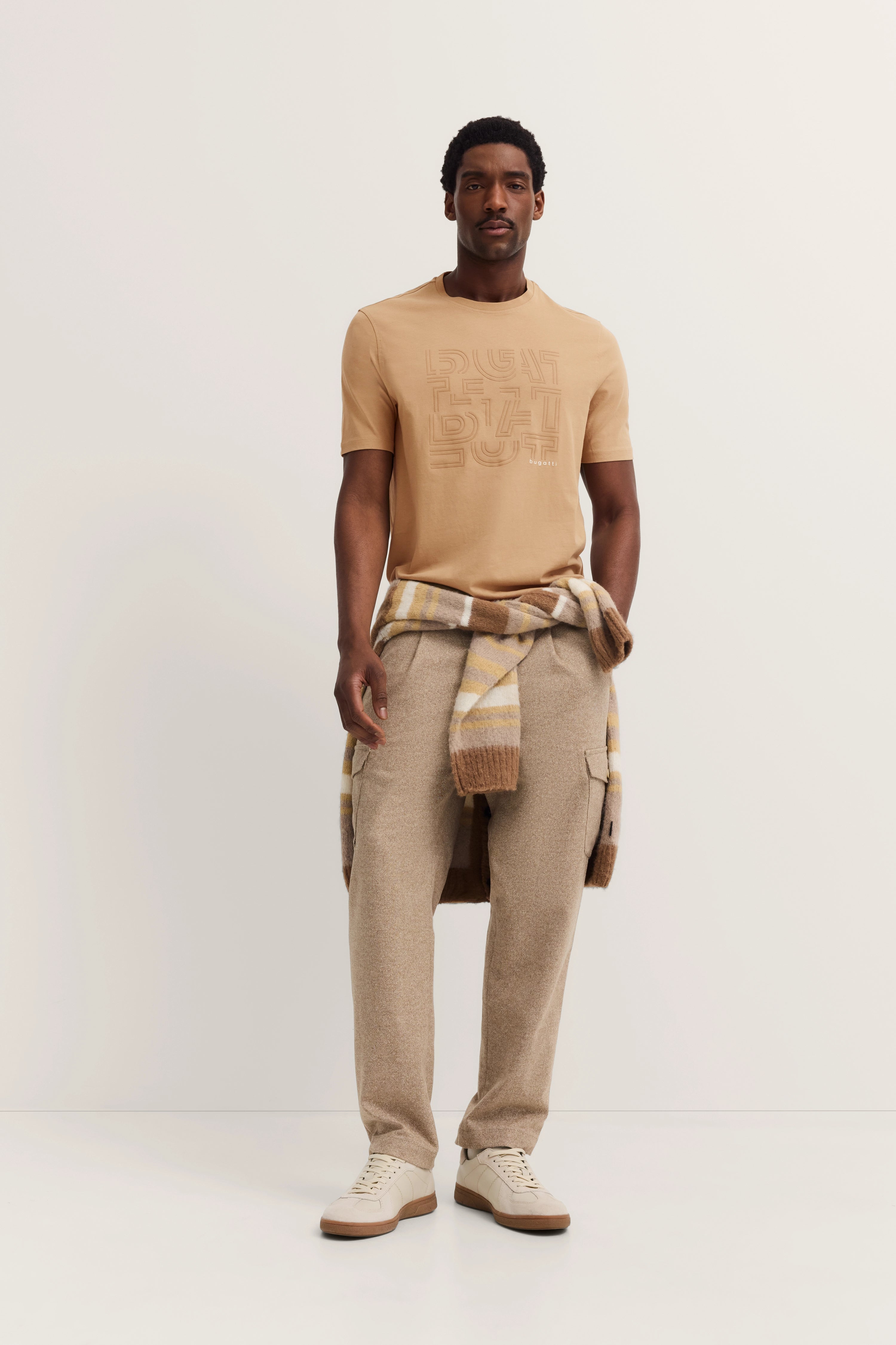 Elegantes bugatti Modeloutfit mit beigem Shirt, kariertem Schal und Hose in Beige, lässig posierend.