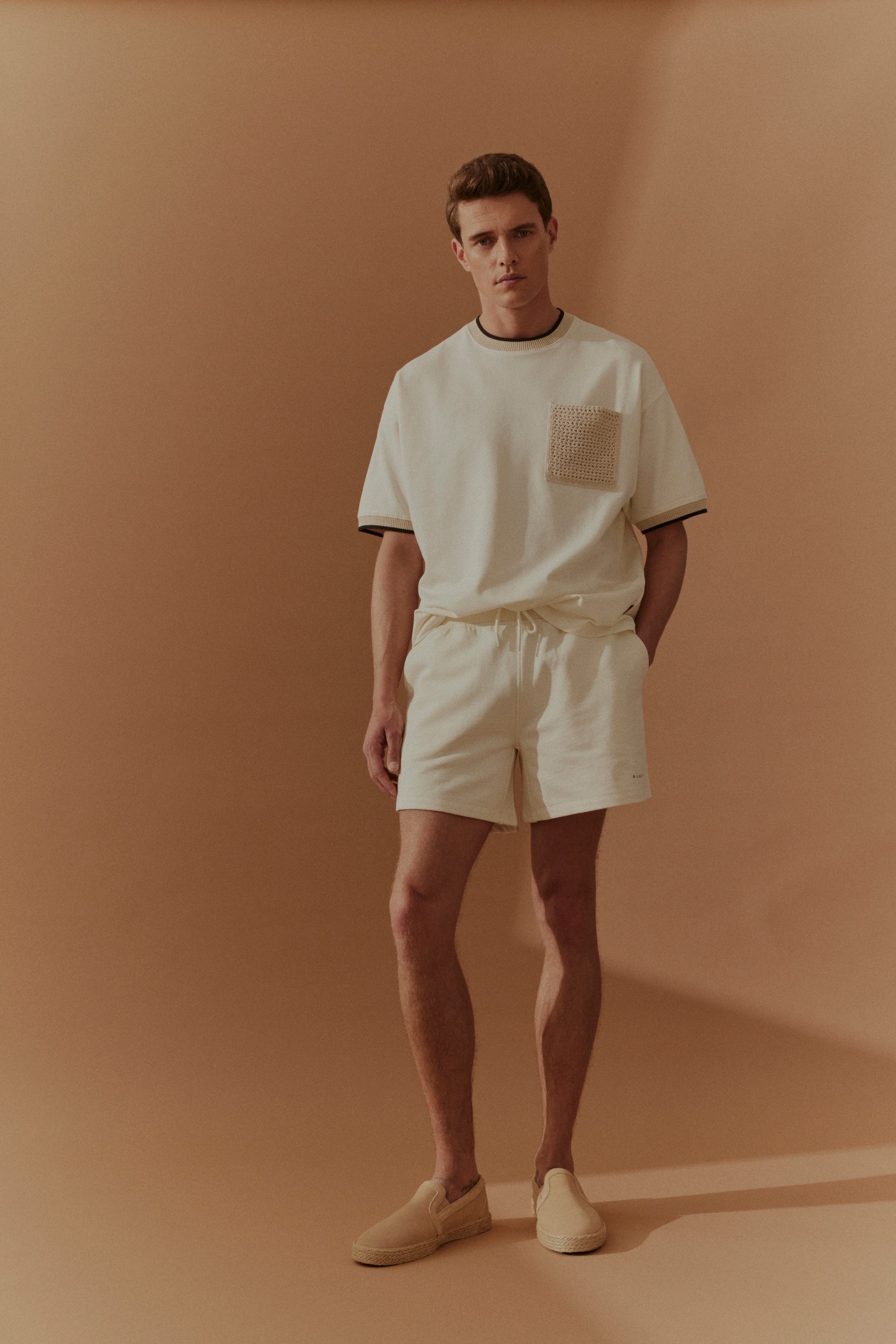 Model in cremefarbenem bugatti T-Shirt und Shorts, legeres Posing vor neutralem Hintergrund.