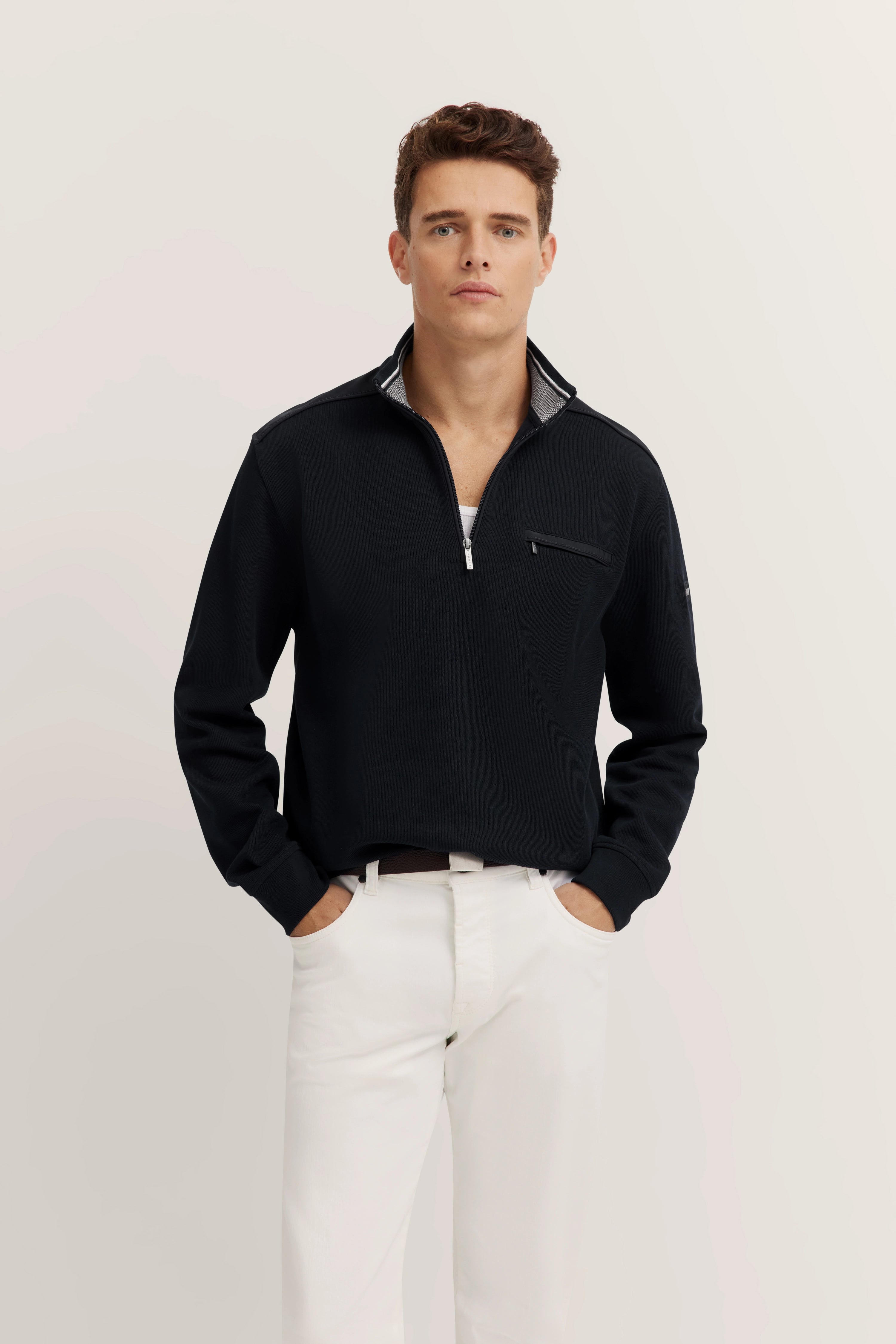 Model in schwarzem bugatti Pullover, weißer Hose, lässige Pose, Hände in den Taschen.
