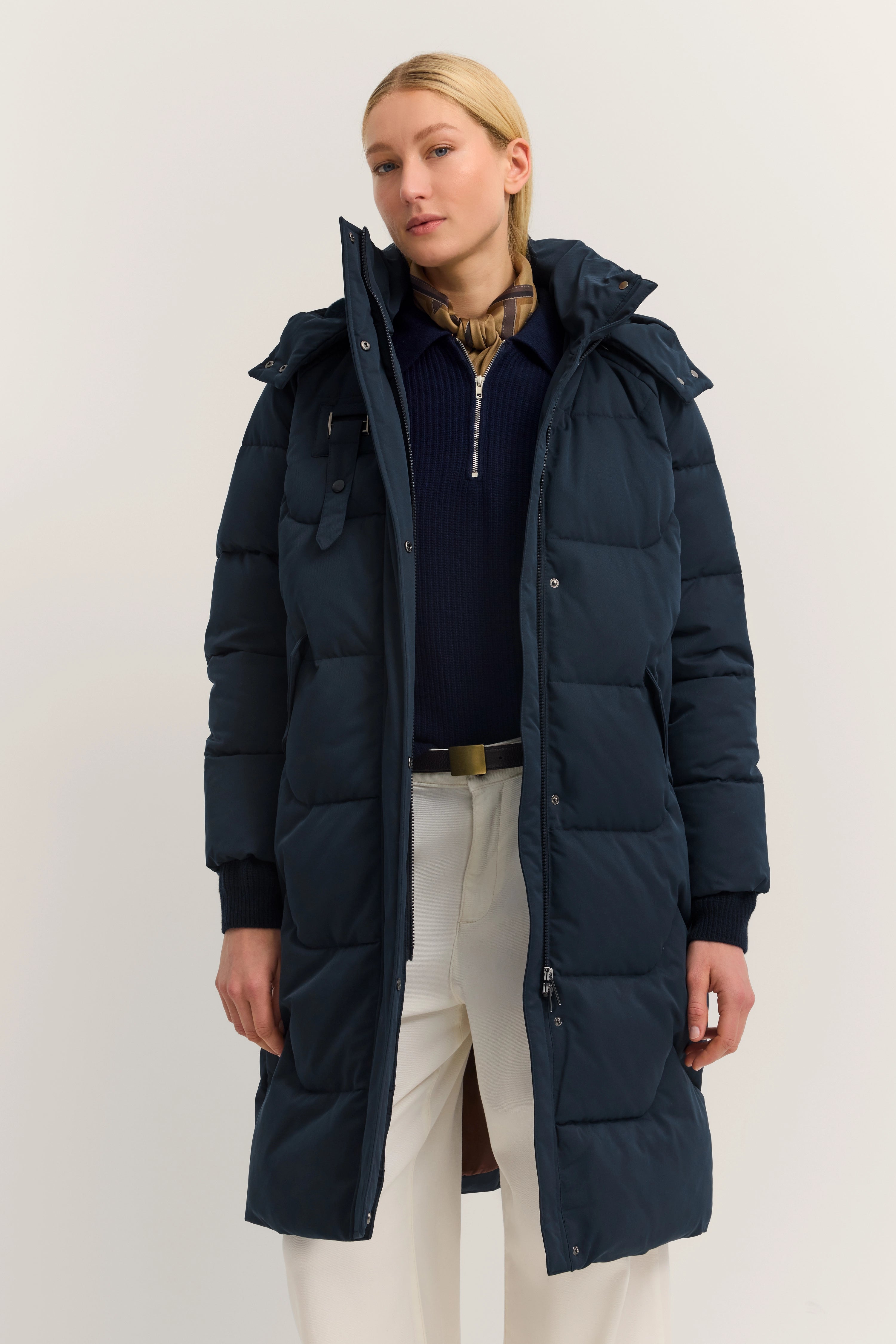 Model in lange, donkerblauwe bugatti donsjas, met beige sjaal en witte broek, staat ontspannen.