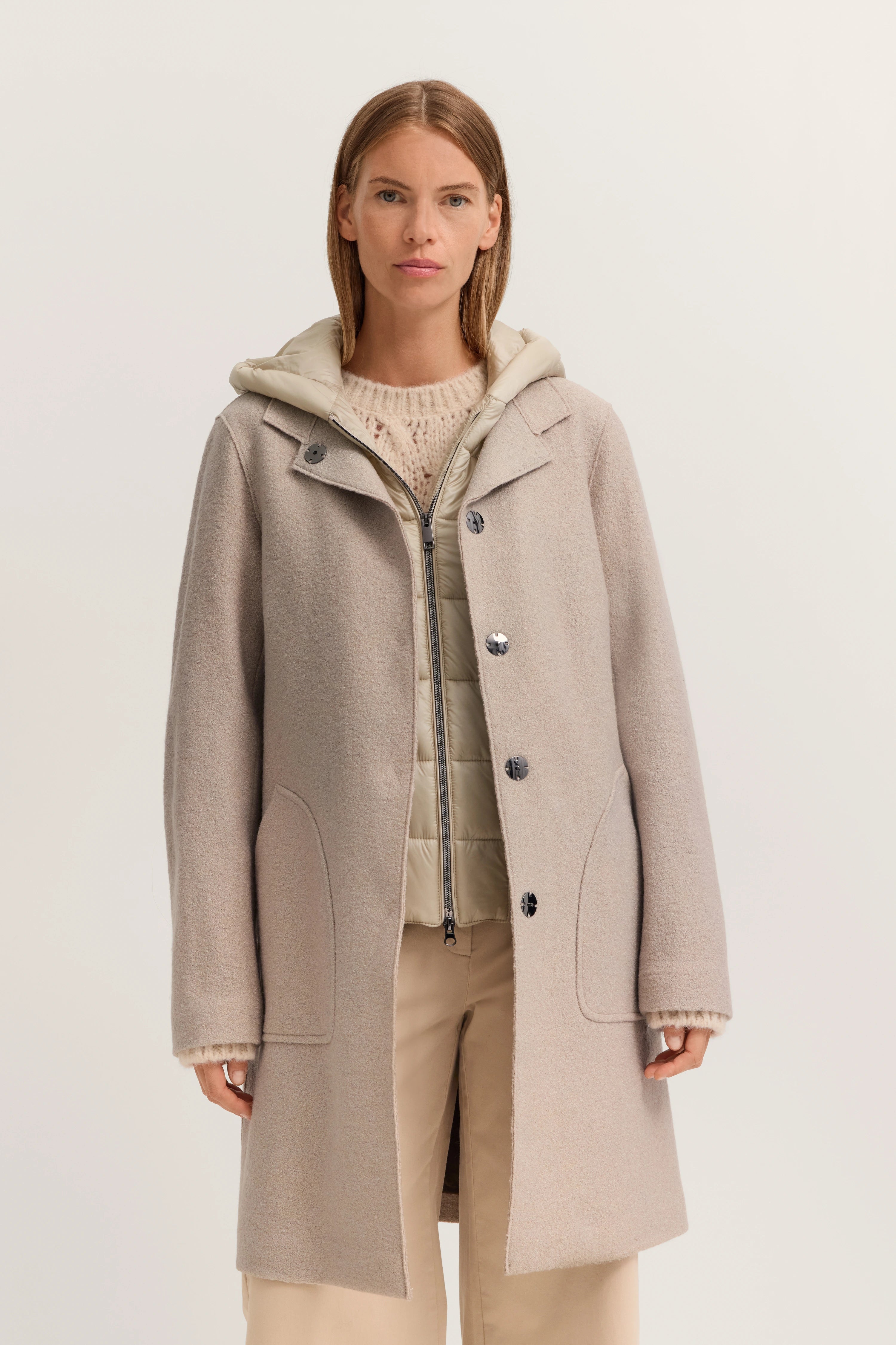 Model in beigem bugatti Mantel, cremefarbener Pullover und Steppjacke. Frontale, entspannte Pose.