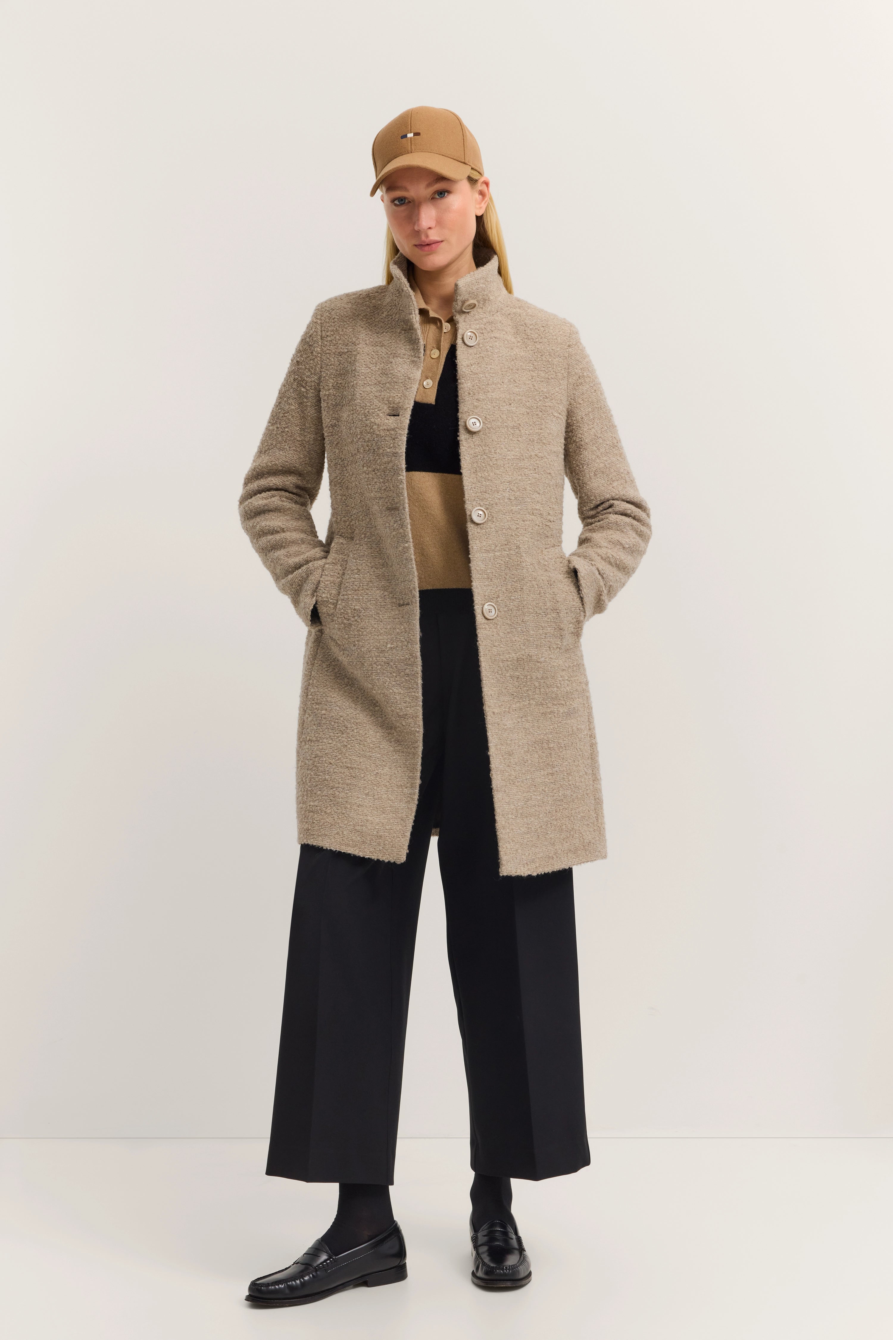Model in beige bugatti jas, zwarte broek, bruine polo en muts, handen in zakken.