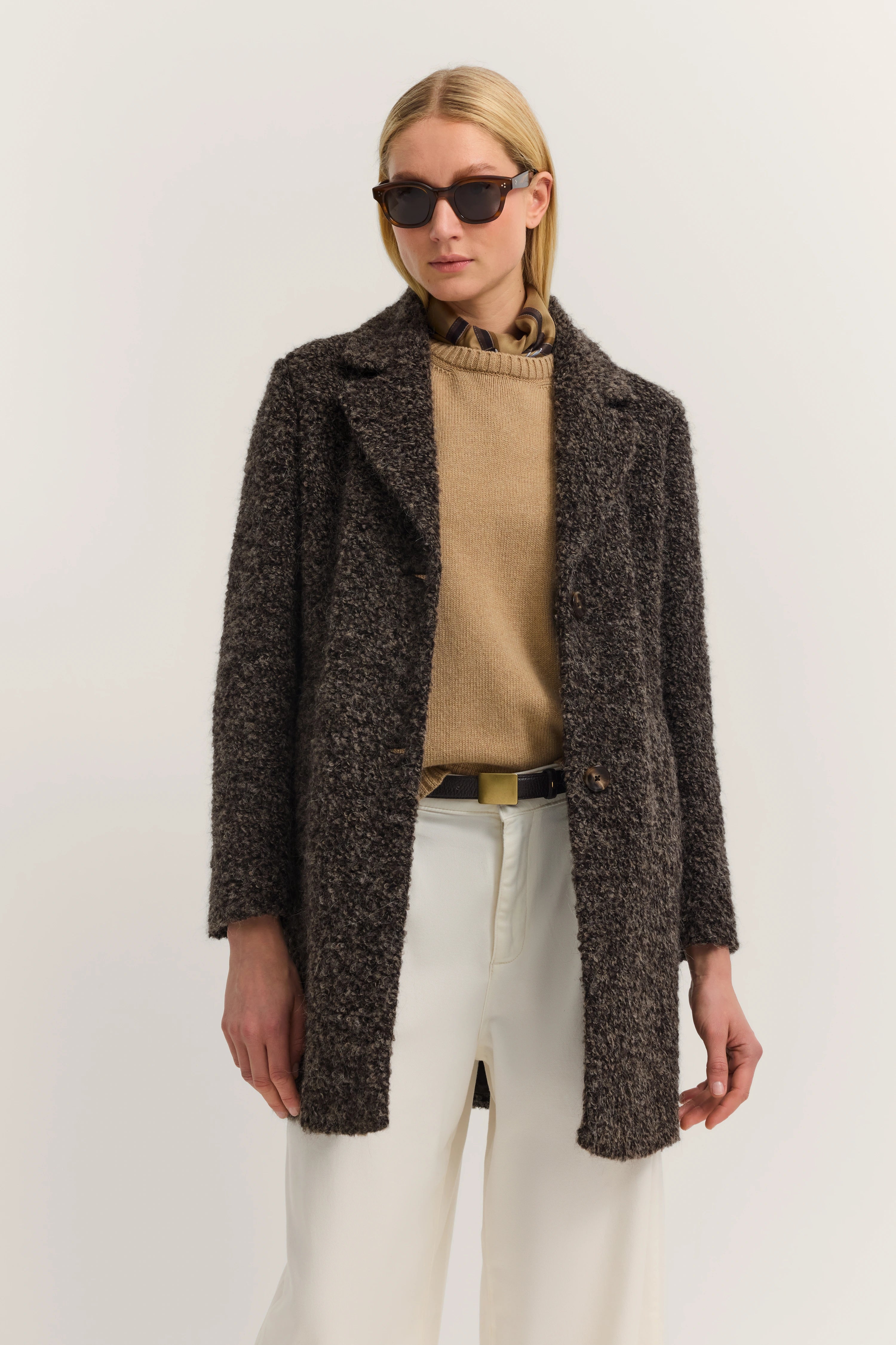 Elegant model in beige trui, grijze jas en witte broek. Nonchalant poserend, bugatti stijl.