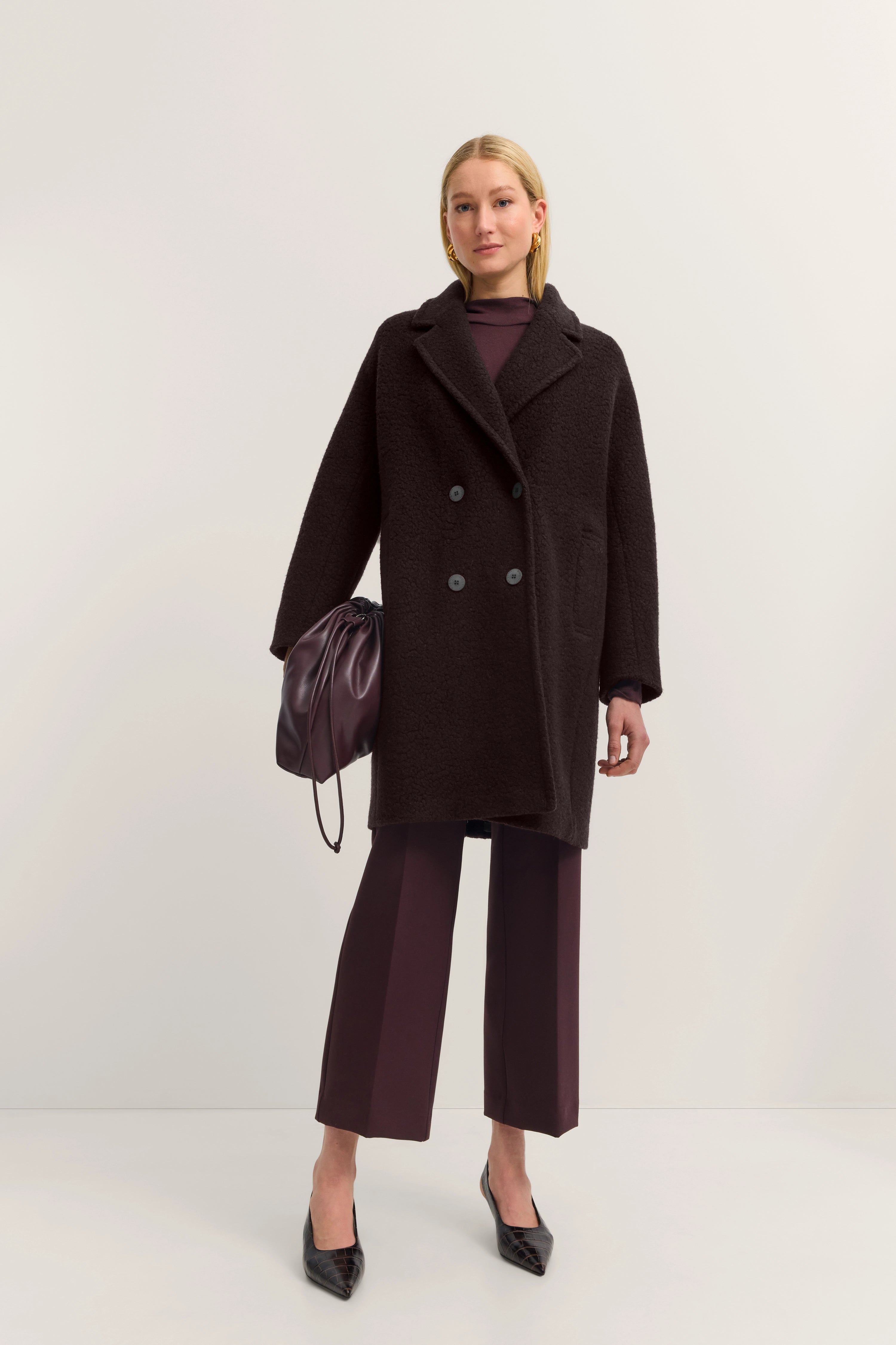 Model in donkerbruine bugatti mantel, bordeauxrode outfit, zwarte schoenen, ontspannen pose.