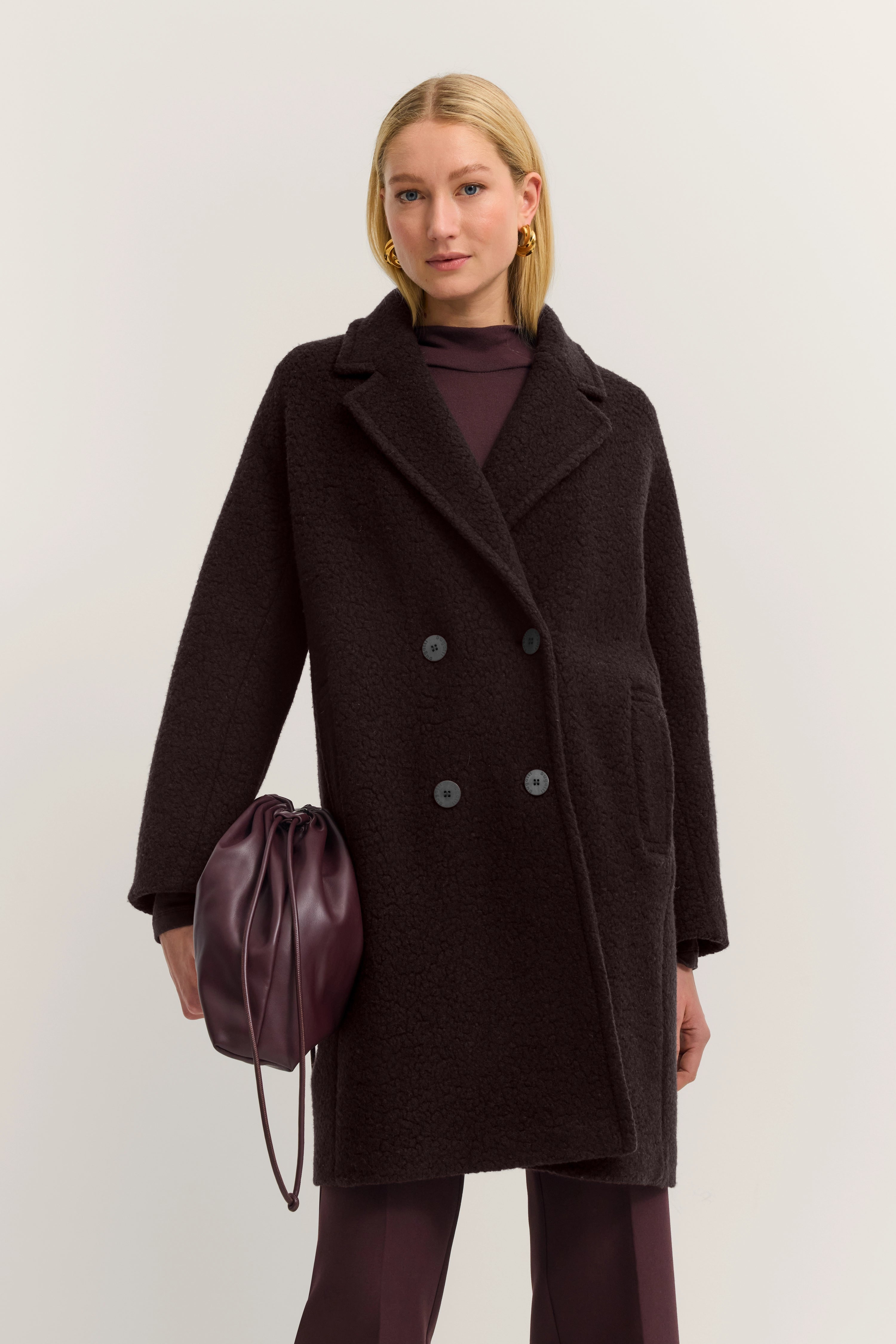 Model in donkerbruine bugatti mantel, wijnrode broek, bijpassende tas, ontspannen pose.