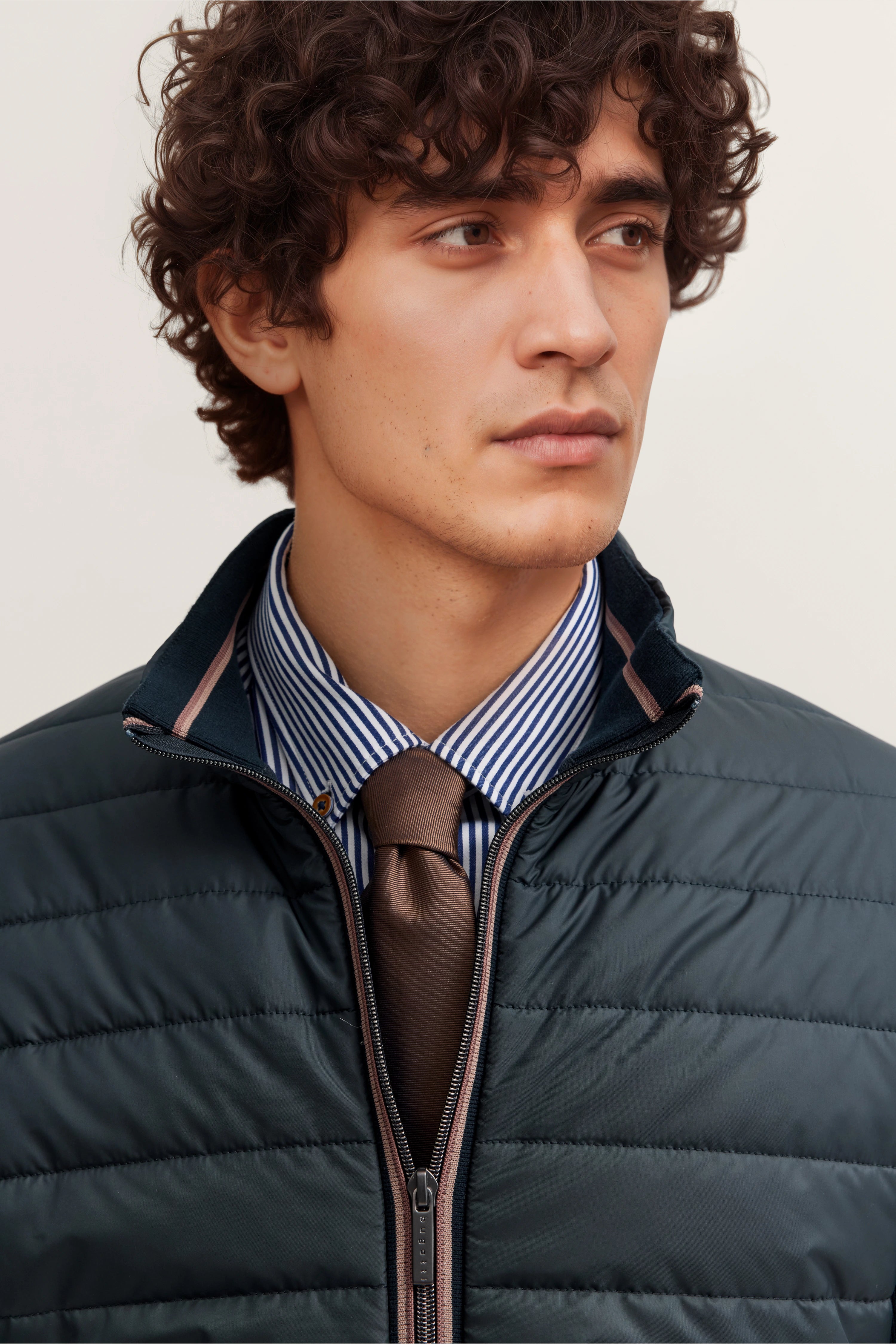 bugatti Model in dunkelblauer Steppjacke, gestreiftem Hemd und brauner Krawatte, schaut ernst zur Seite.