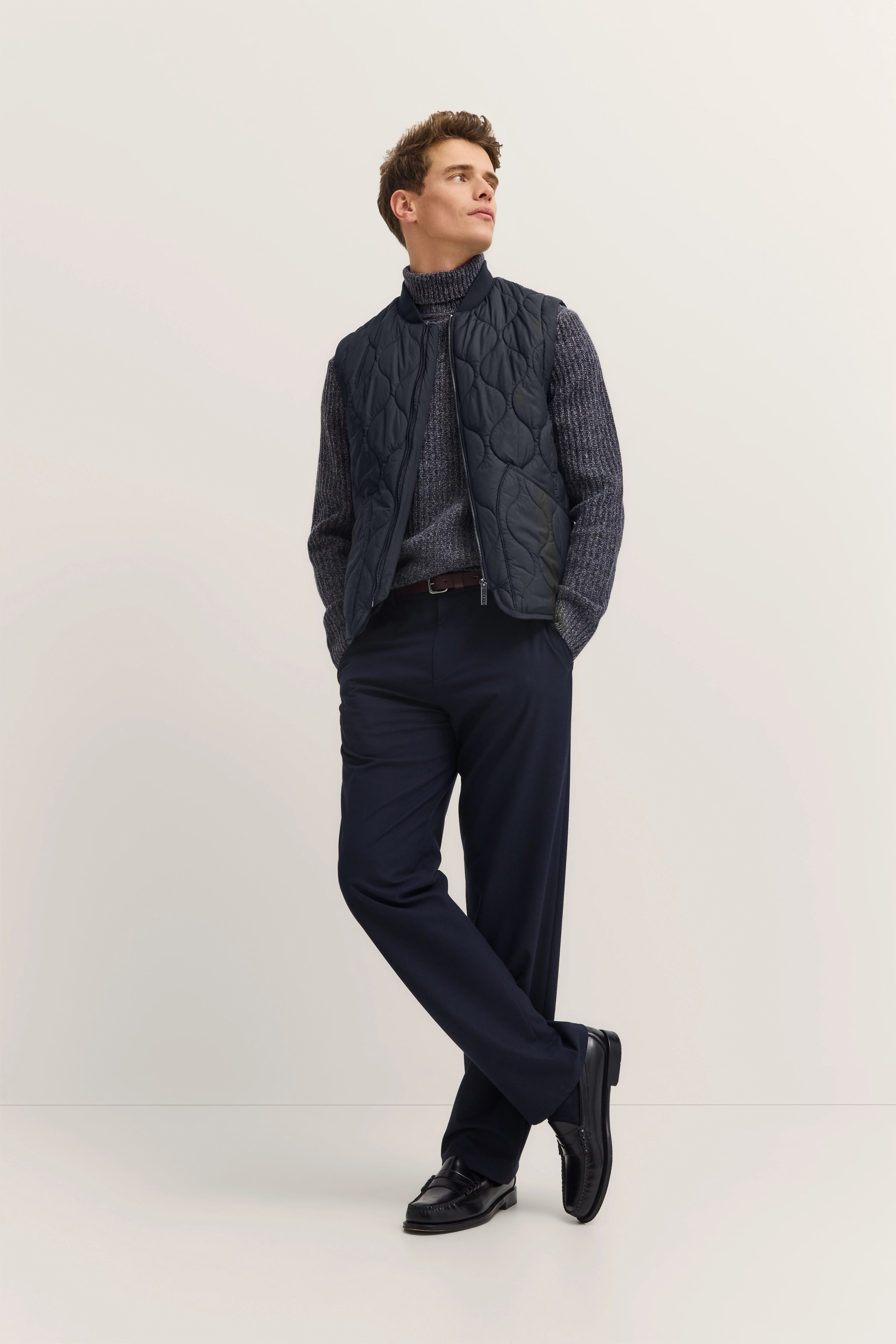 Model in gray turtleneck, quilted vest, navy pants, casually posing.