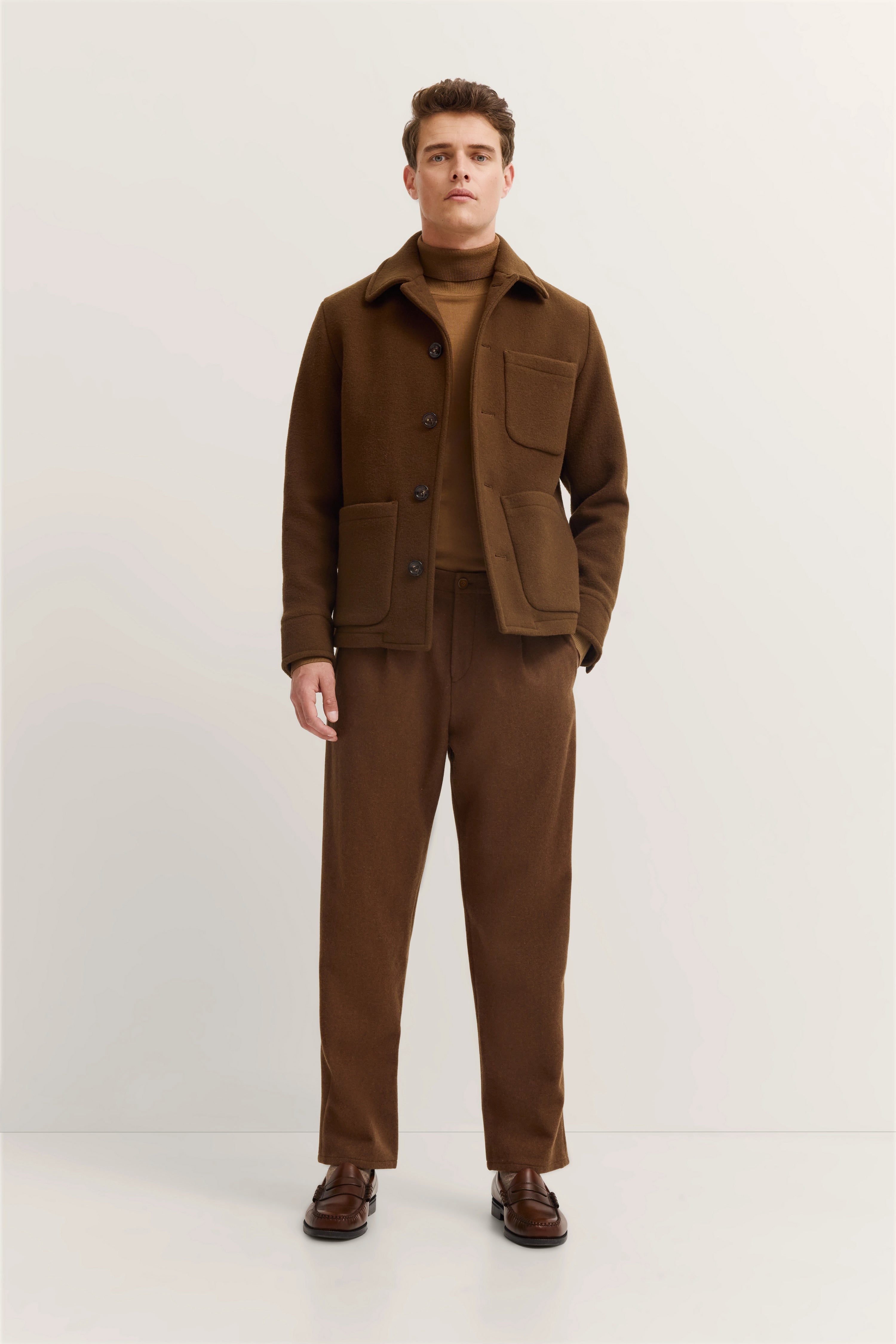 Alttext: Model in braunem bugatti-Outfit, Mantel mit Taschen, Rollkragenpullover, gerade Hose, entspannte Pose.