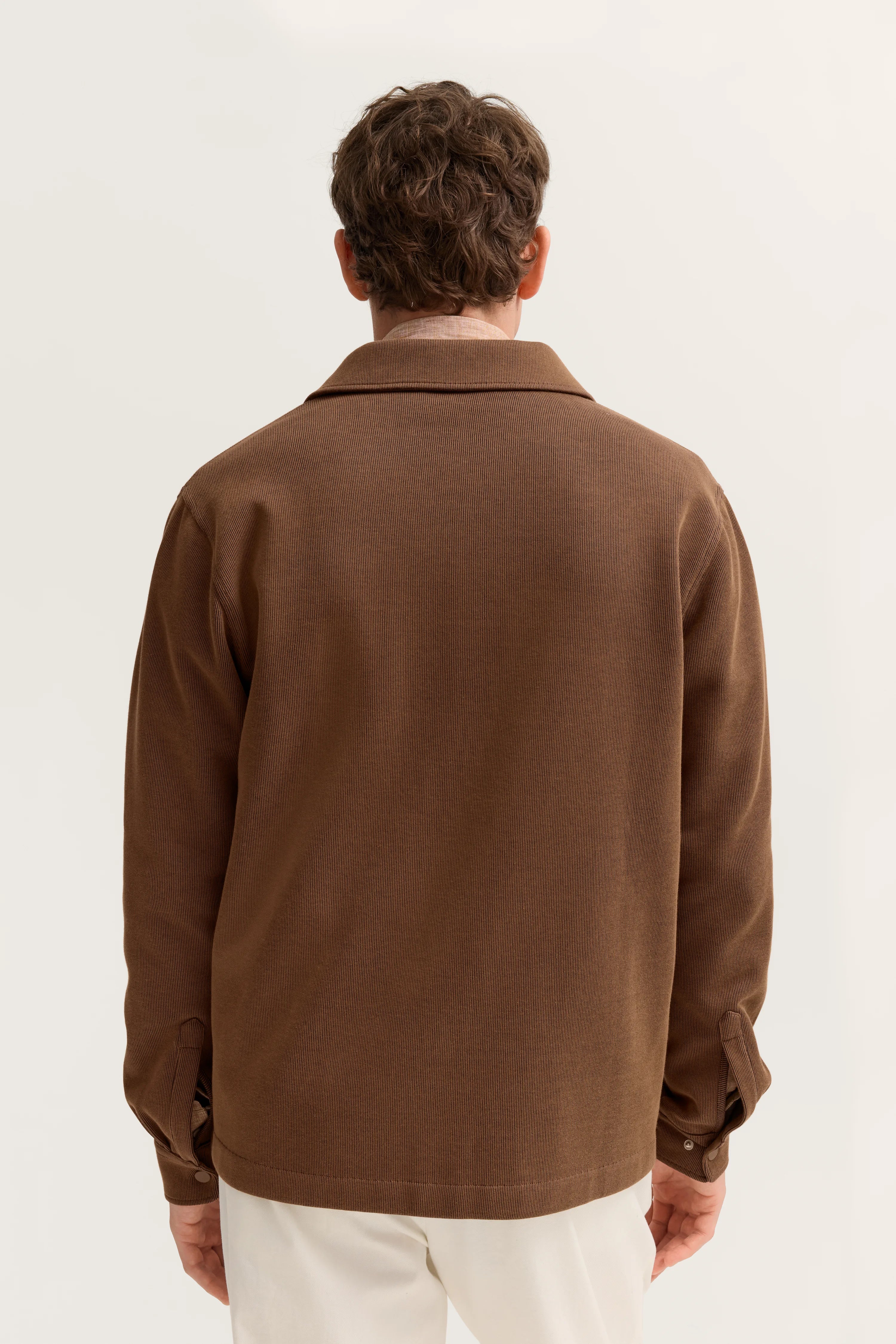 Braune Jacke von hinten, legeres Modelbild, entspannte Pose, kombiniert mit weißer Hose.