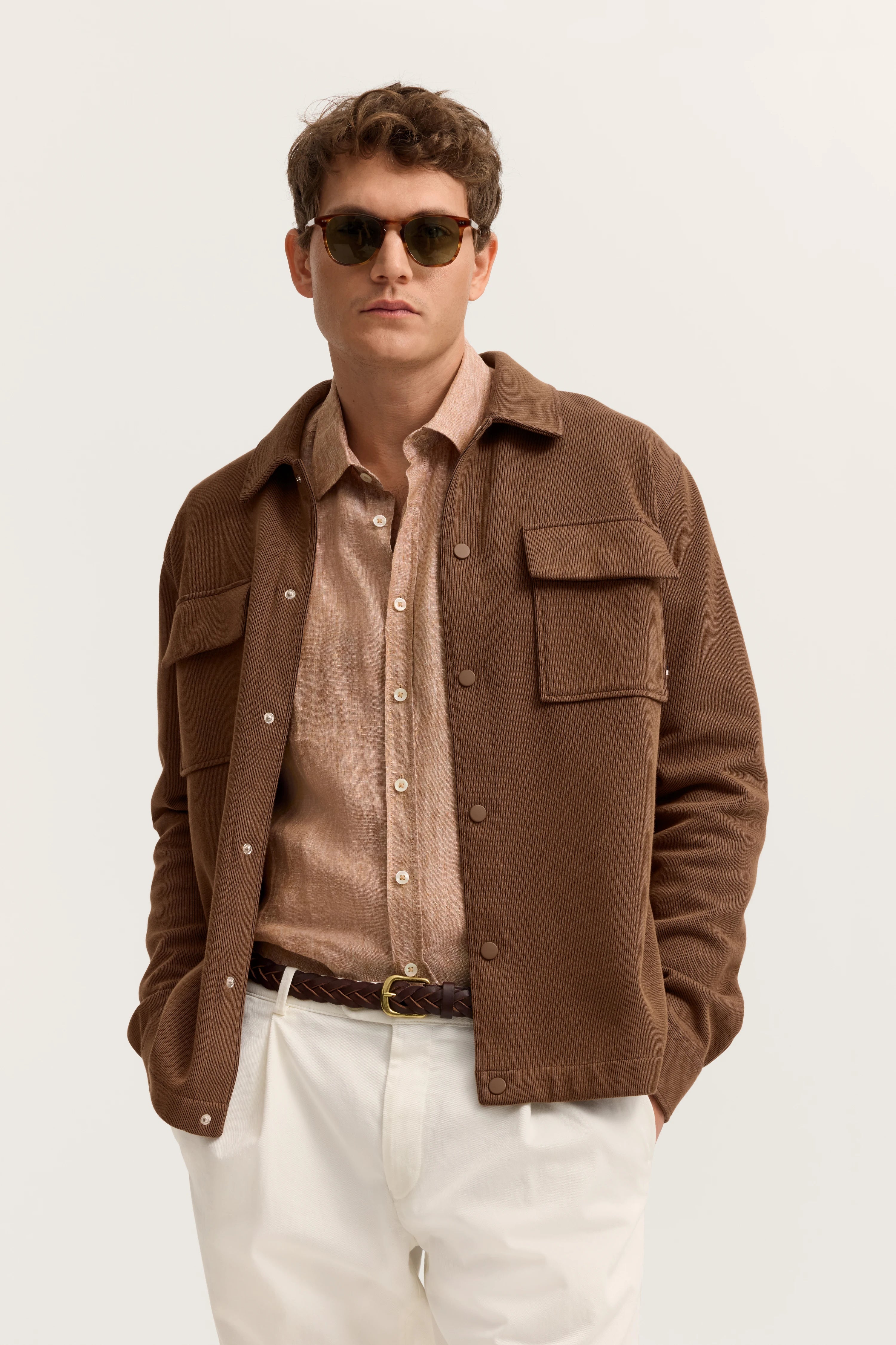 Bugatti Modelbild: Beige Hemd, braune Jacke, weiße Hose. Lässig stehend, Hände in Hosentaschen.