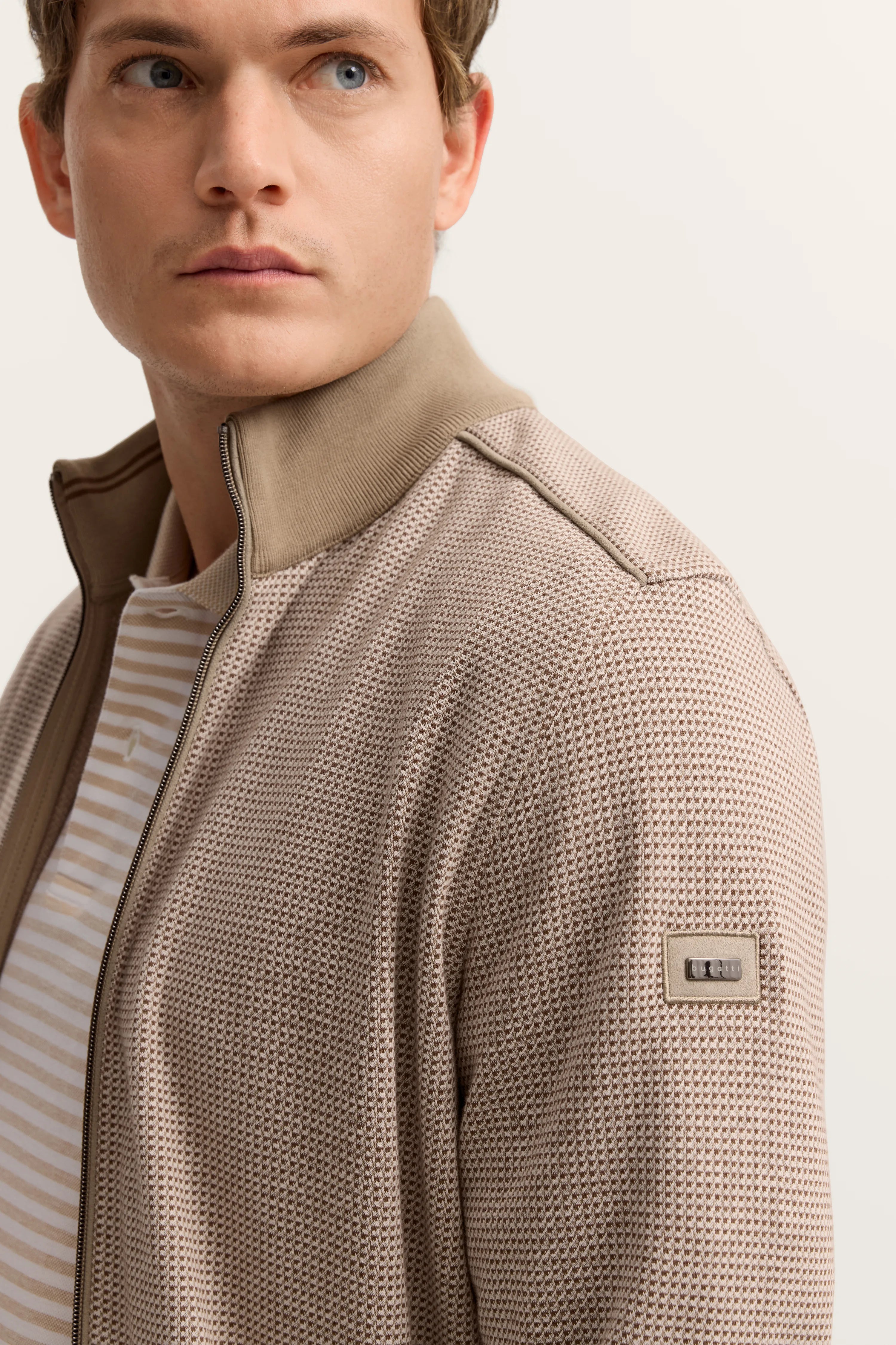 Rückenansicht Modell in beige gemusterter bugatti-Jacke und weißen Hosen, leger stehend.