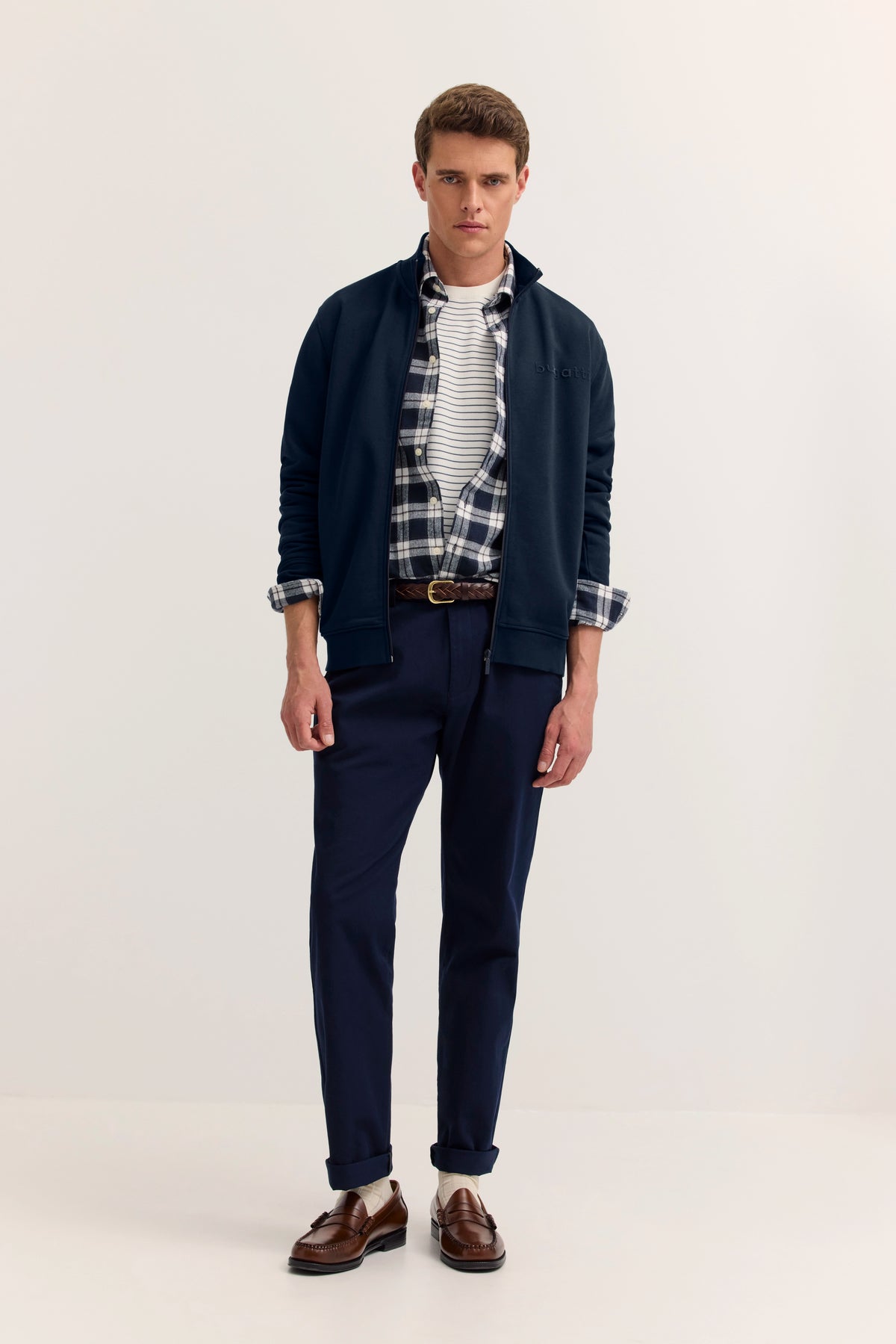 Model in bugatti-Outfit: Dunkelblaue Jacke, kariertes Hemd, gestreiftes Shirt, blaue Hose, braune Schuhe.