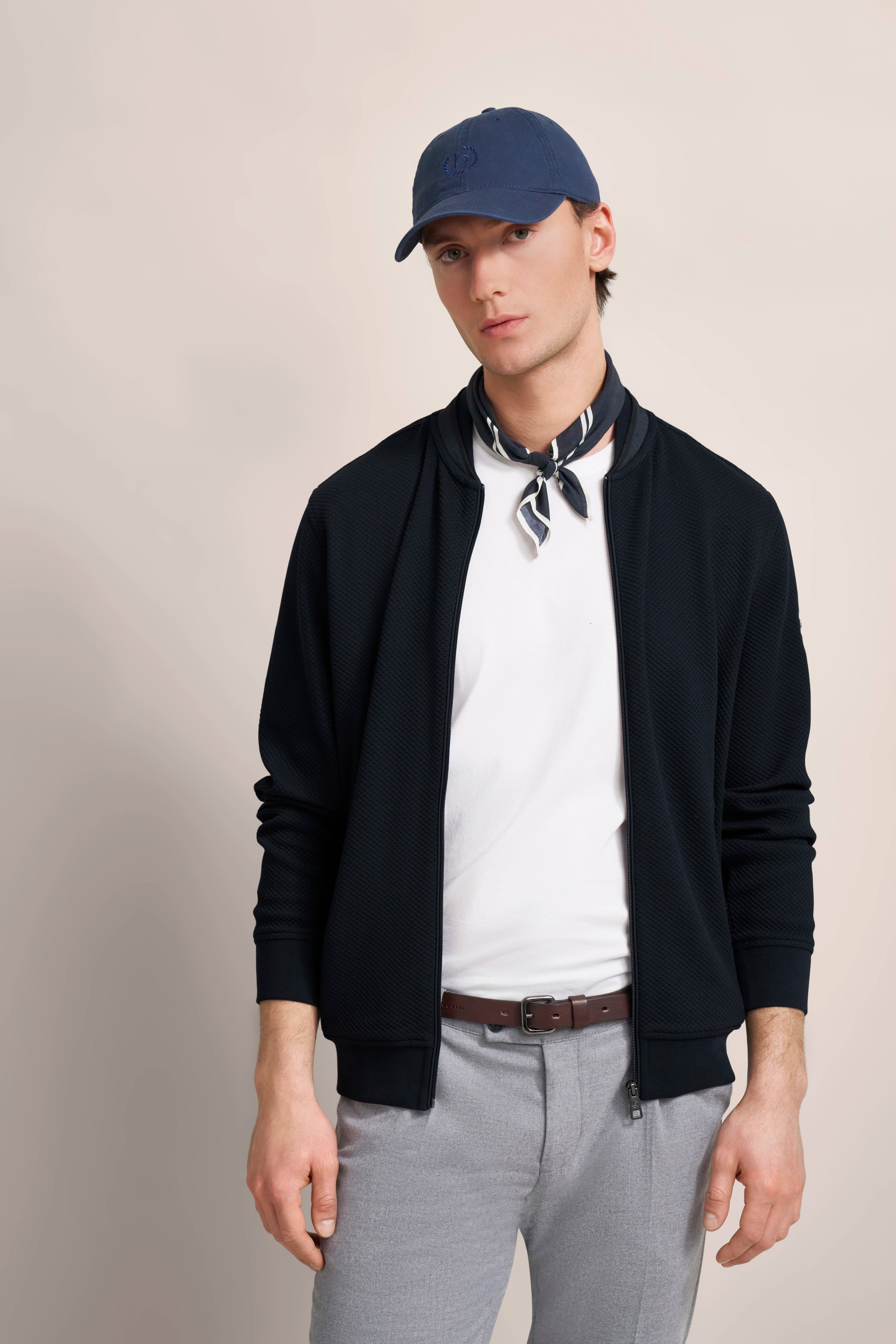 Model in bugatti outfit: dark jacket, white shirt, gray pants, blue cap, scarf. Relaxed pose.