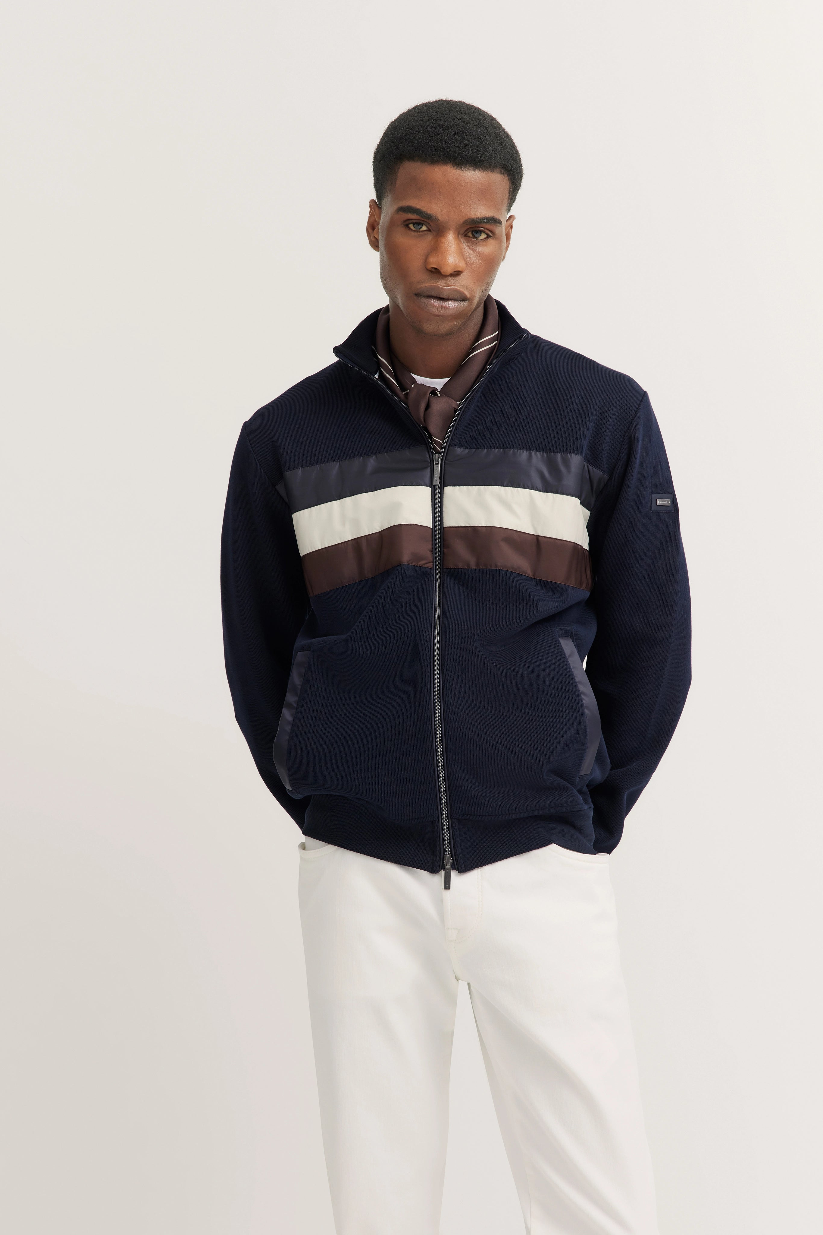 Model in bugatti jacket with blue, white, brown stripes, paired with white pants, standing casually.