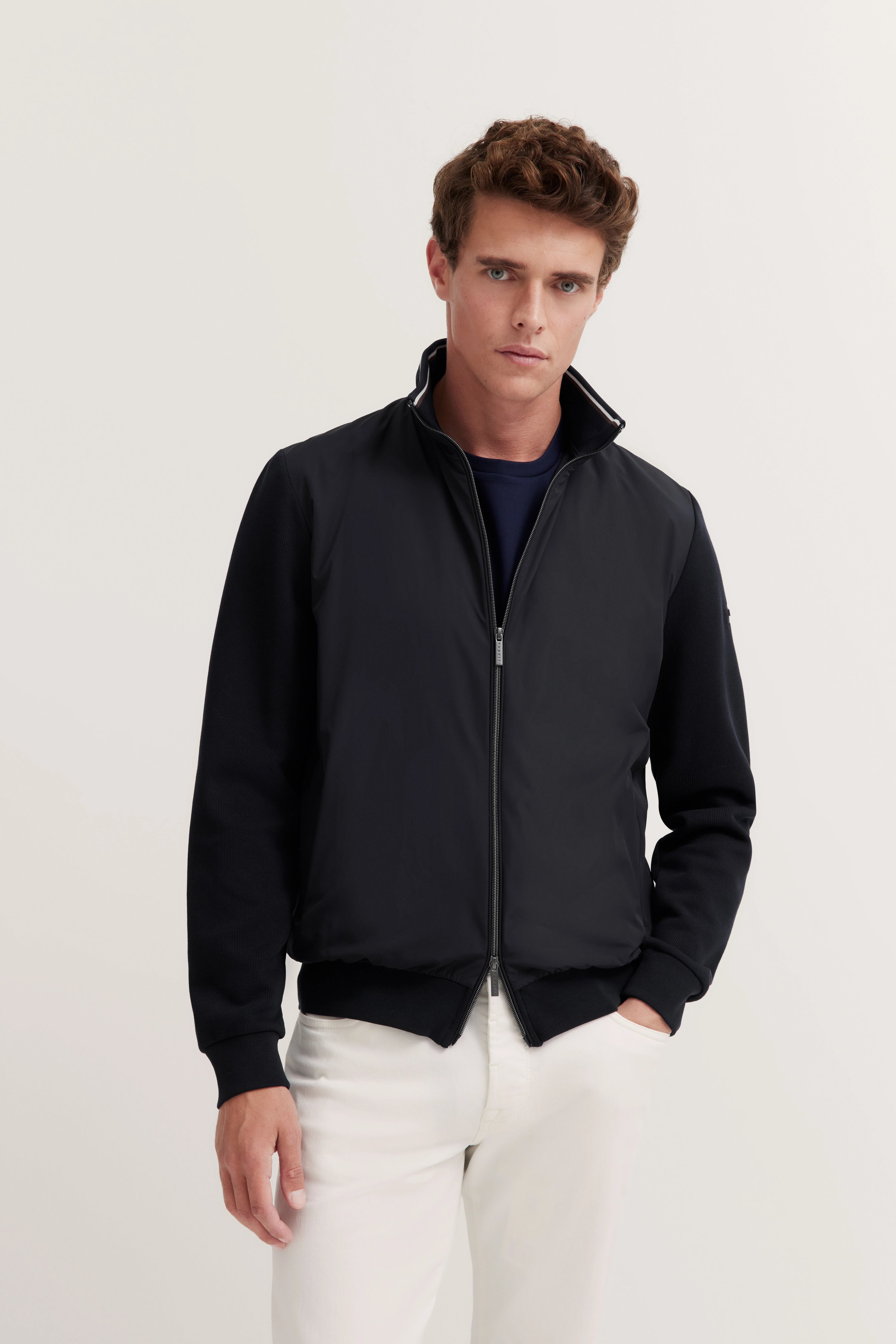 Model in schwarzer bugatti Jacke und weißer Hose, leger stehend, mit entspanntem Ausdruck.