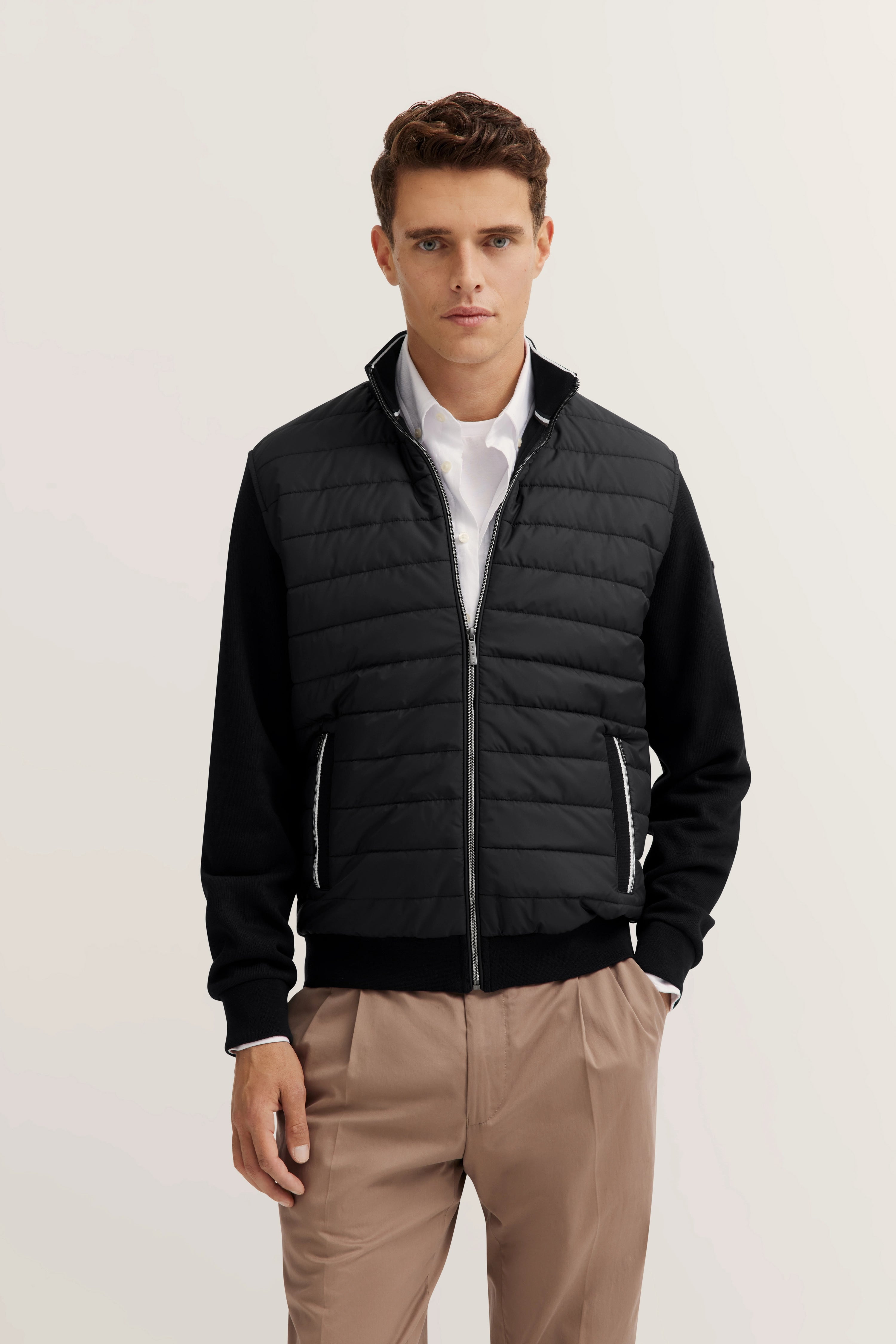 Model in schwarzer bugatti Steppjacke, weißem Hemd und beigen Hosen, frontal stehend.