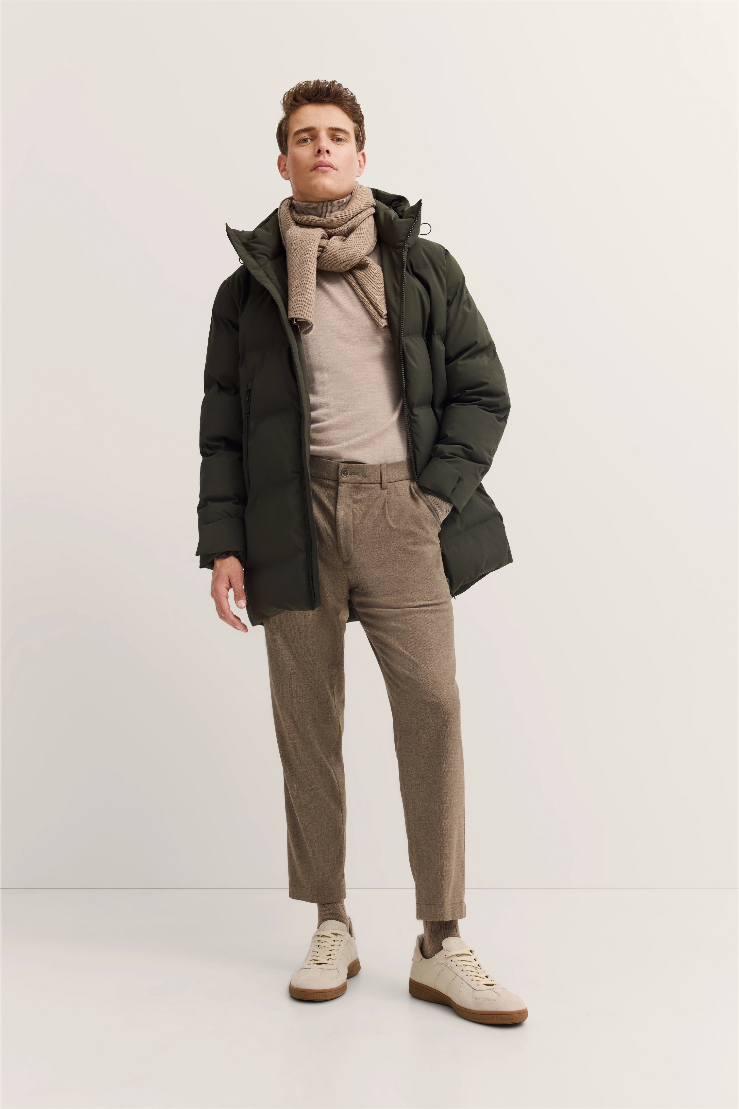 Model im legeren Outfit: olivgrüne Daunenjacke, beigefarbener Schal, helles Shirt, braune Hose, helle Sneaker.