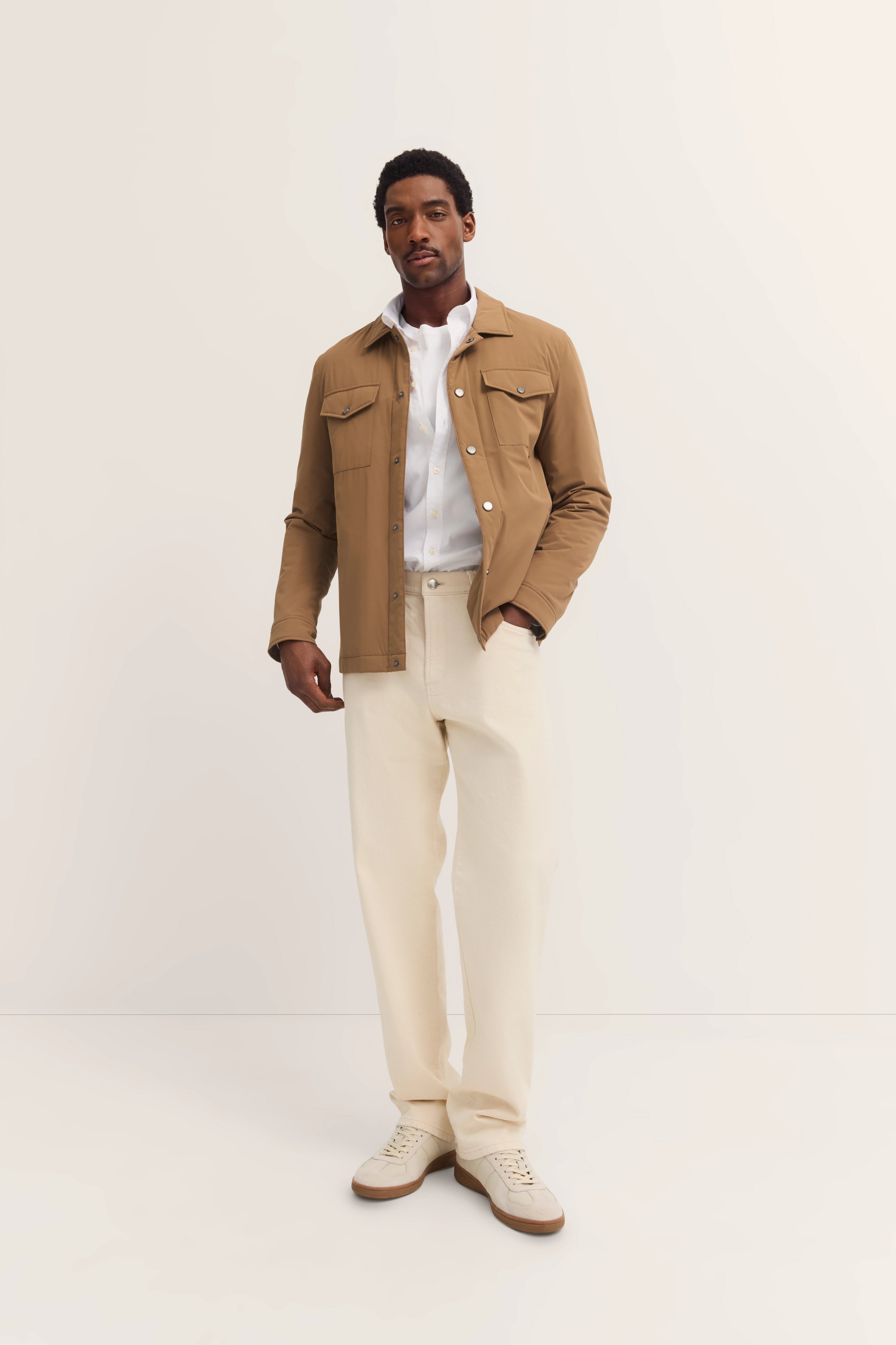 Model draagt bugatti outfit: beige jas, wit hemd, crèmekleurige broek, lichte schoenen. Ontspannen pose.