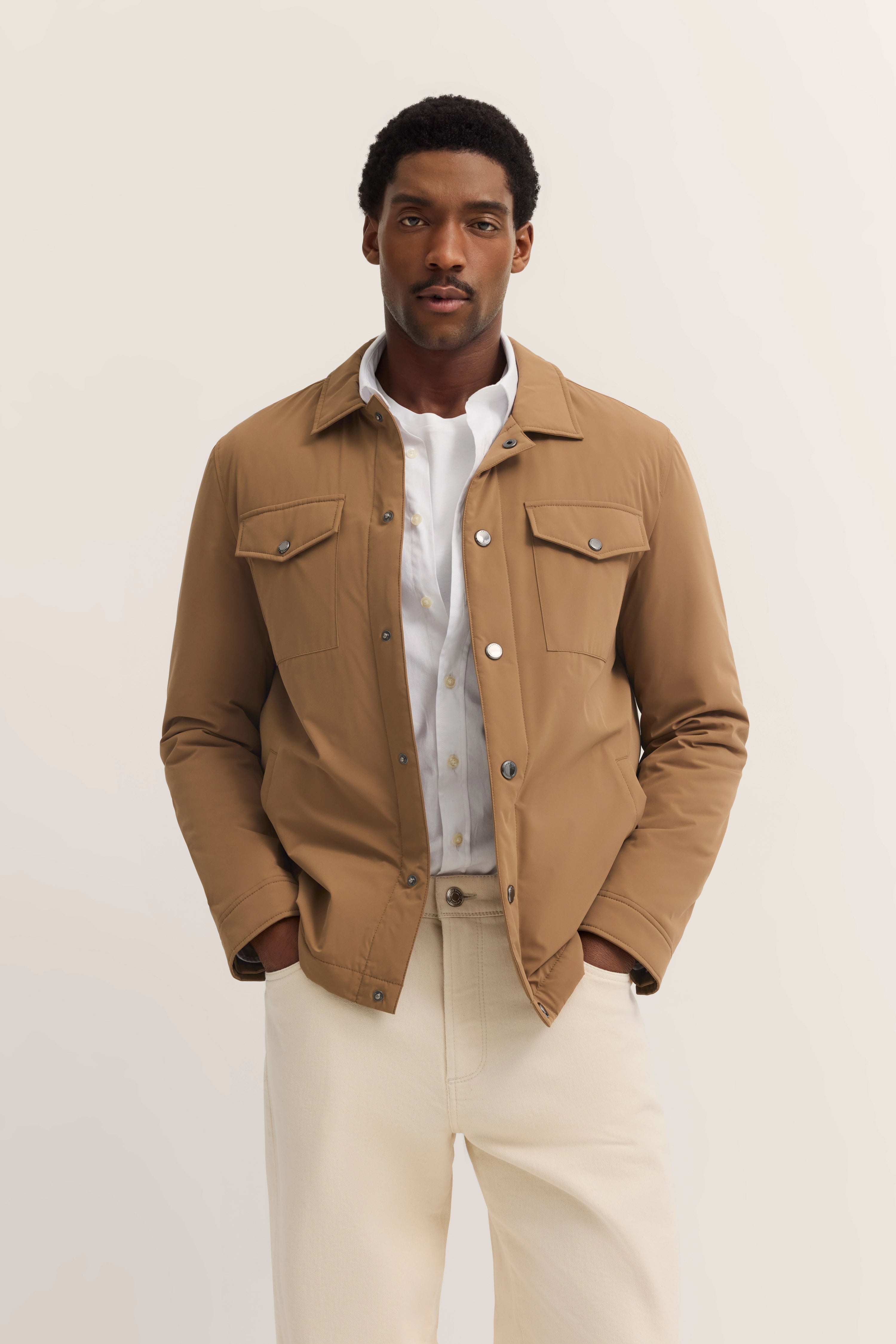 Model in beige jack en wit overhemd, ontspannen houding.