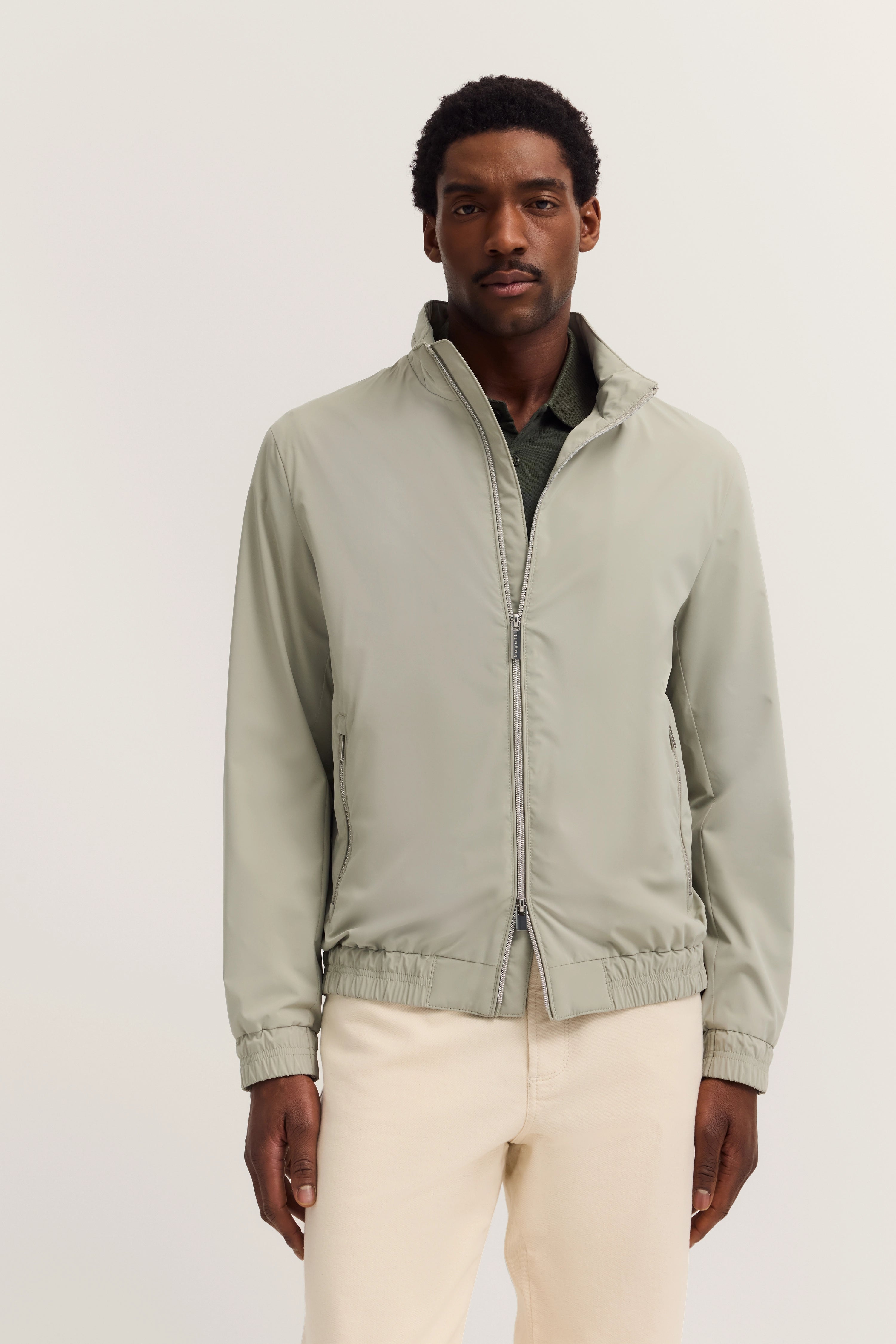 Model draagt bugatti jas in lichtgrijs, gecombineerd met donkergroene blouse en beige broek. Rechte houding.