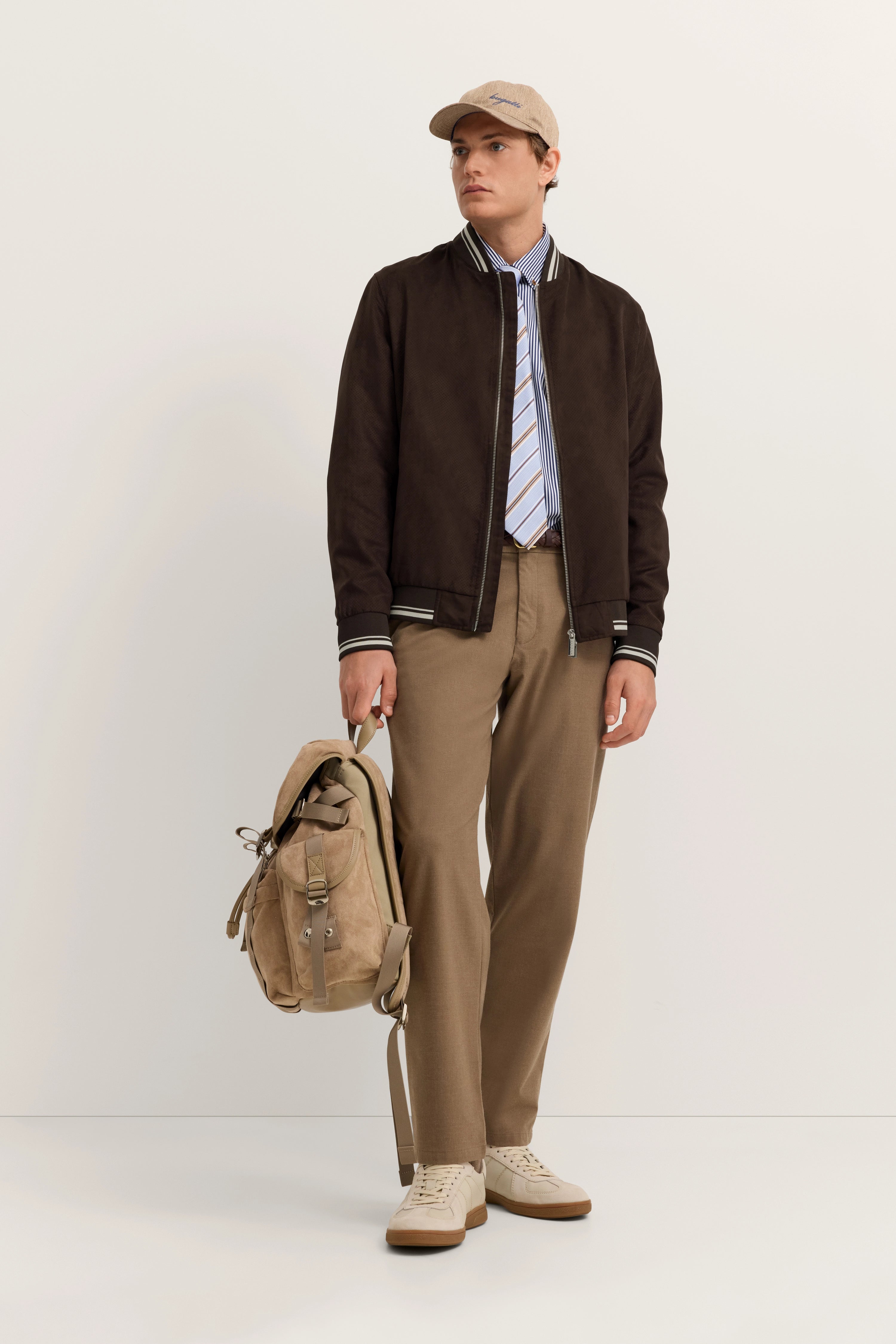 Model in brauner bugatti Jacke, gestreiftem Hemd, Krawatte, beigen Hosen, Sneakers und Cap, casual Pose.