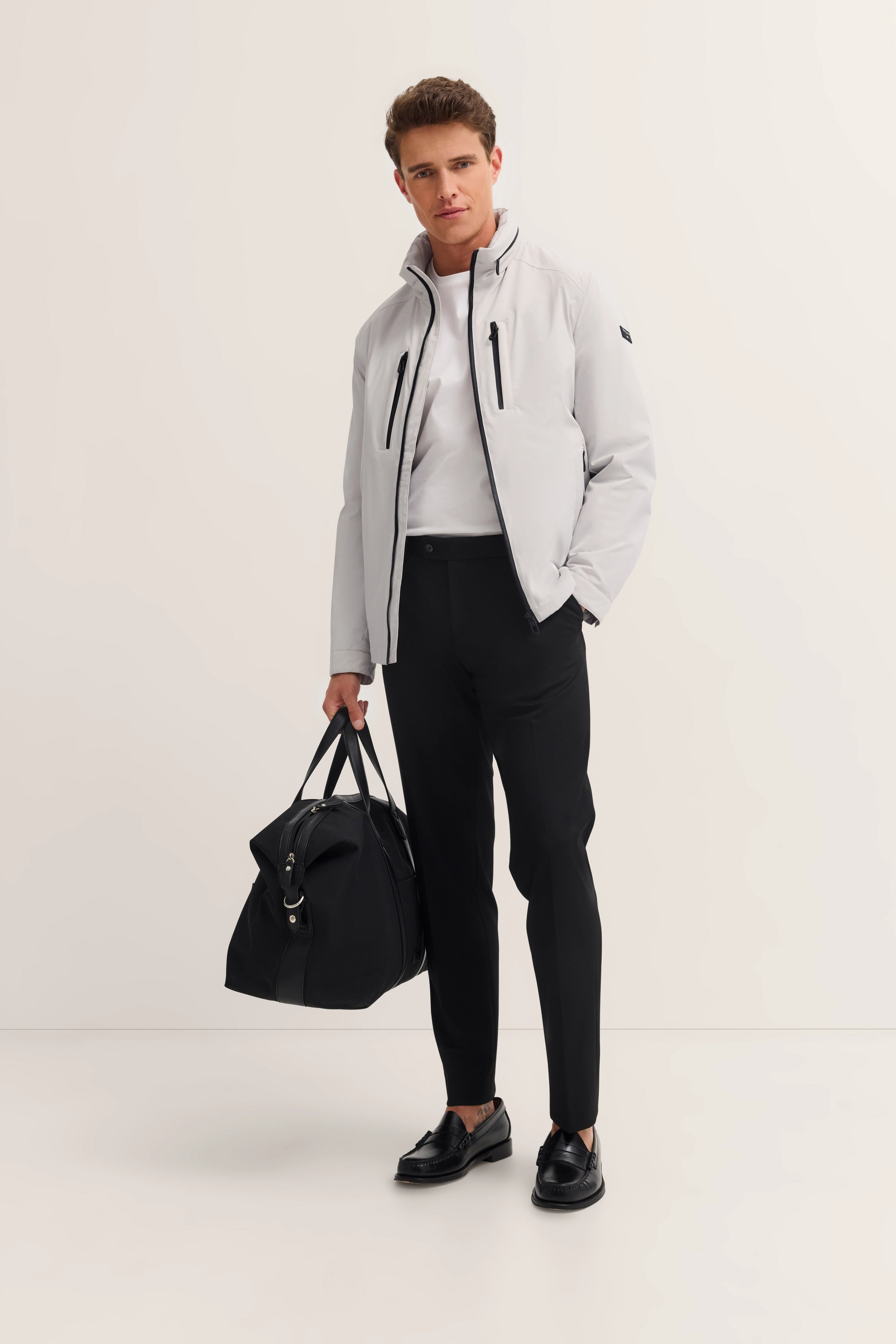 Model in white bugatti jacket, black pants, white shirt, black shoes, carrying black bag.