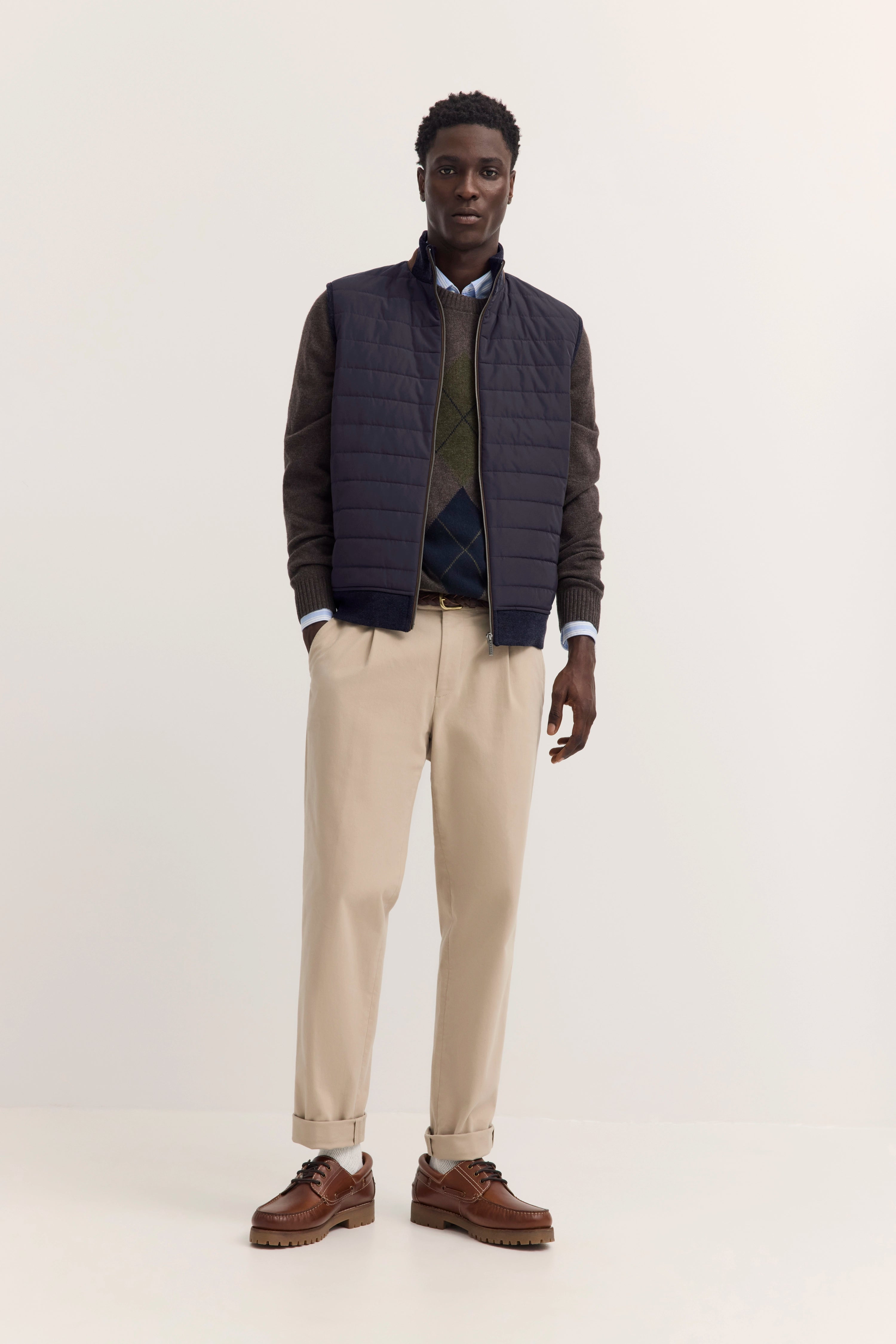 Model in navy Steppweste, braunem Pullover, grüner Argyle-Weste, beigefarbenen Chinos. Elegantes Standposing.