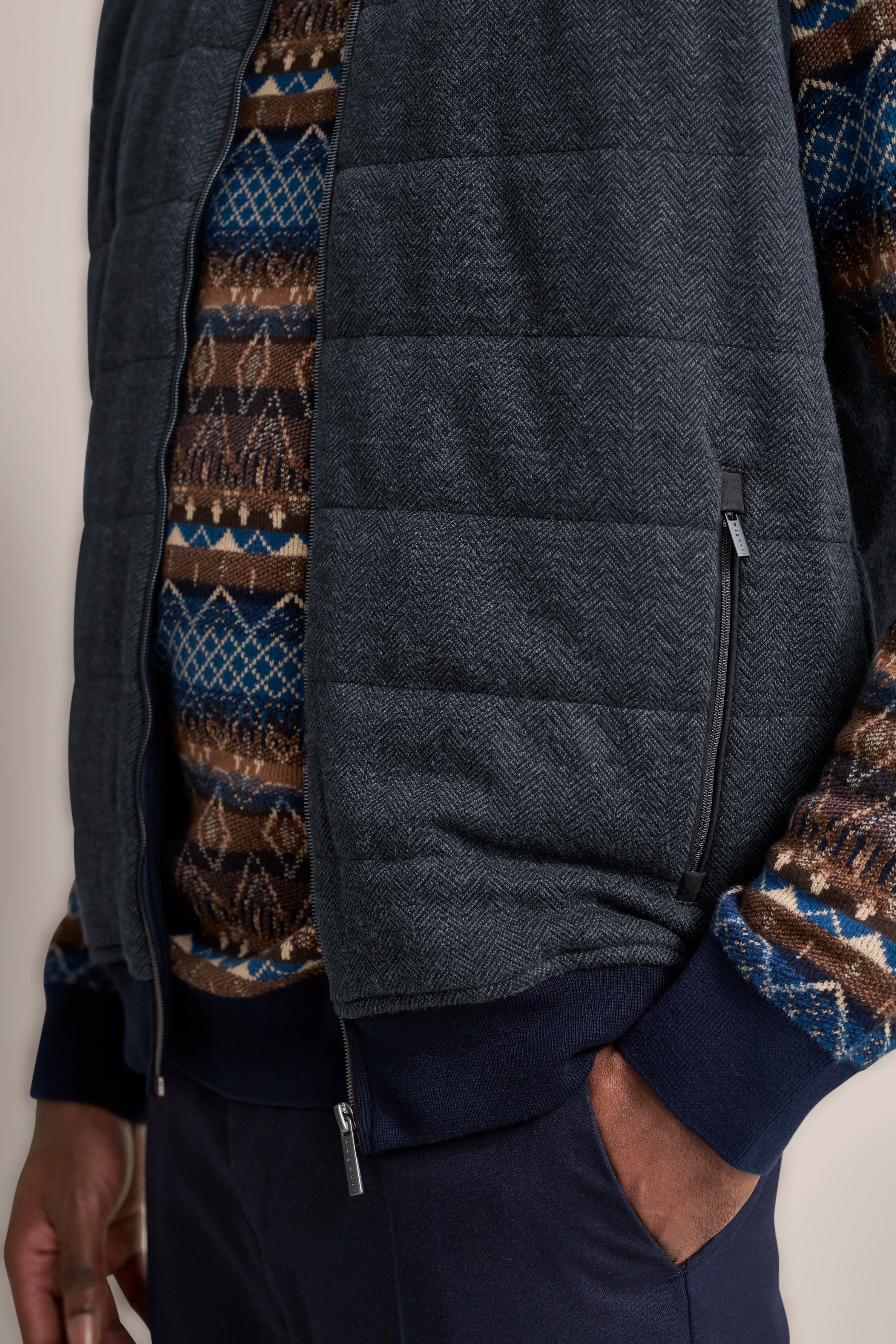 Gesteppte, graue Weste mit gemustertem Pullover in Blau-Braun und dunkelblauer Hose. Büstenbild.