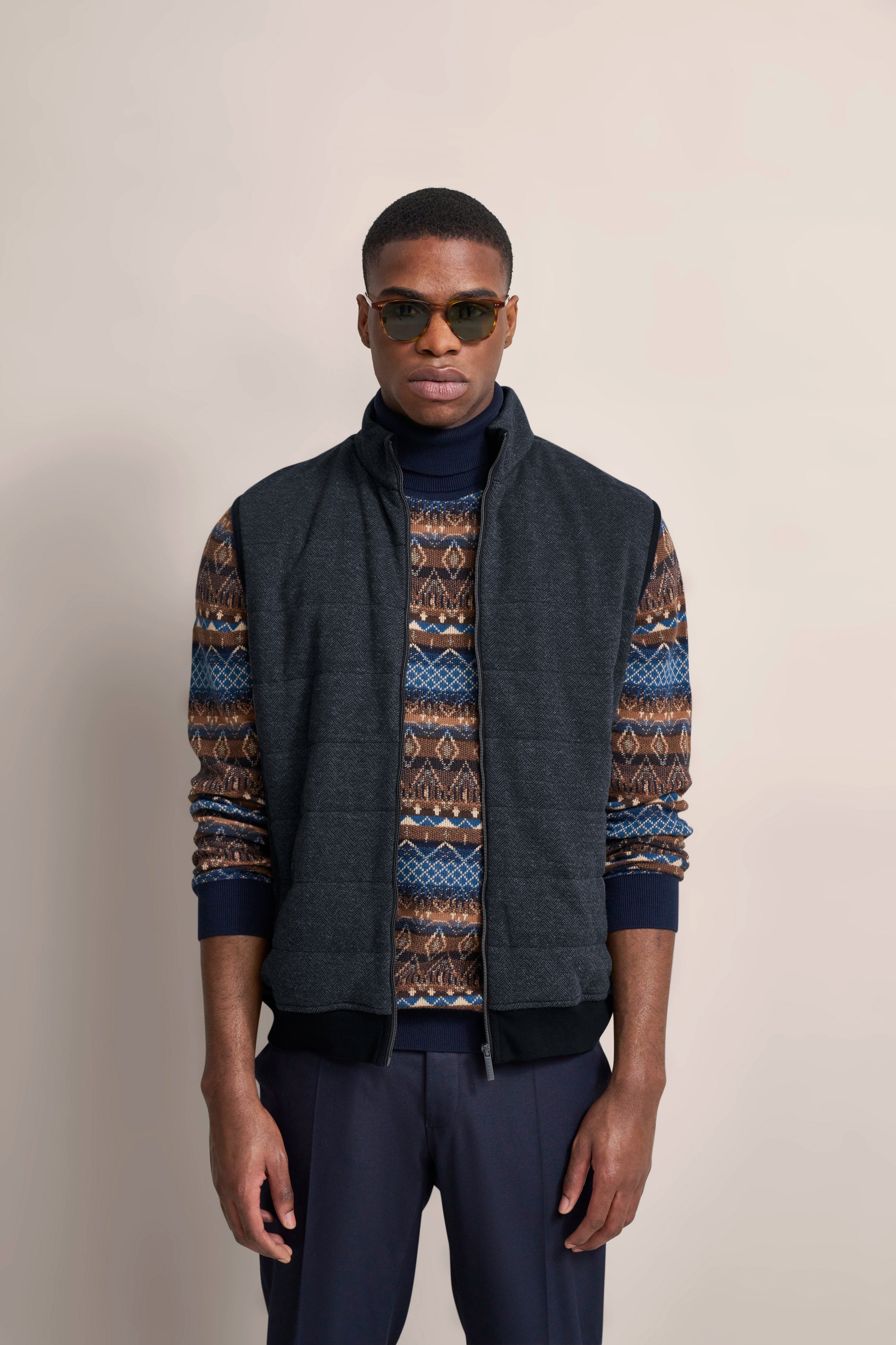 Model mit gemustertem Pullover, dunkler Weste und Hose, frontal stehend, lässig-urbaner Stil.