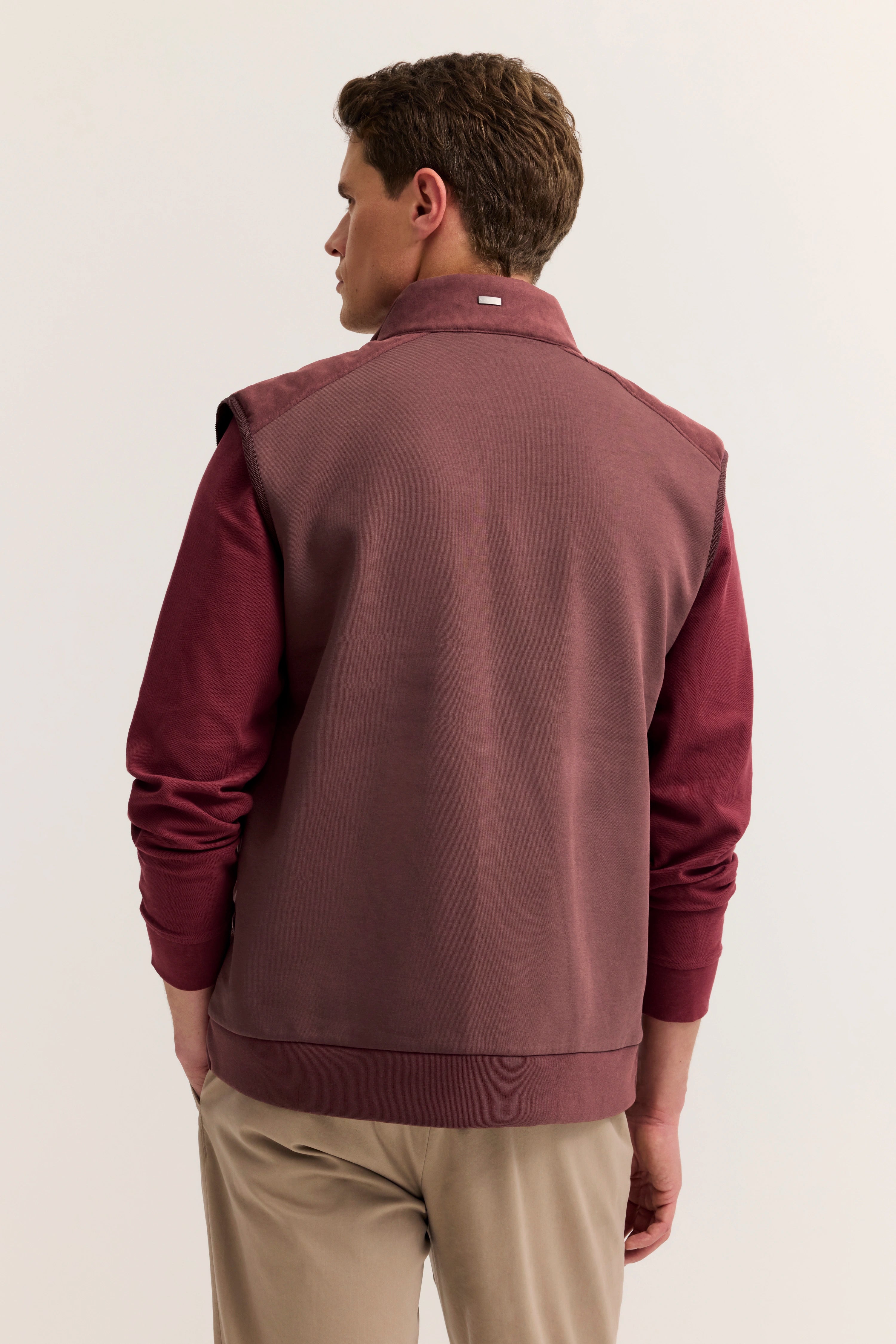 Büstenbild von hinten: Model in bordeauxrotem Pullover und Weste, beige Hose, lässiges Posing.