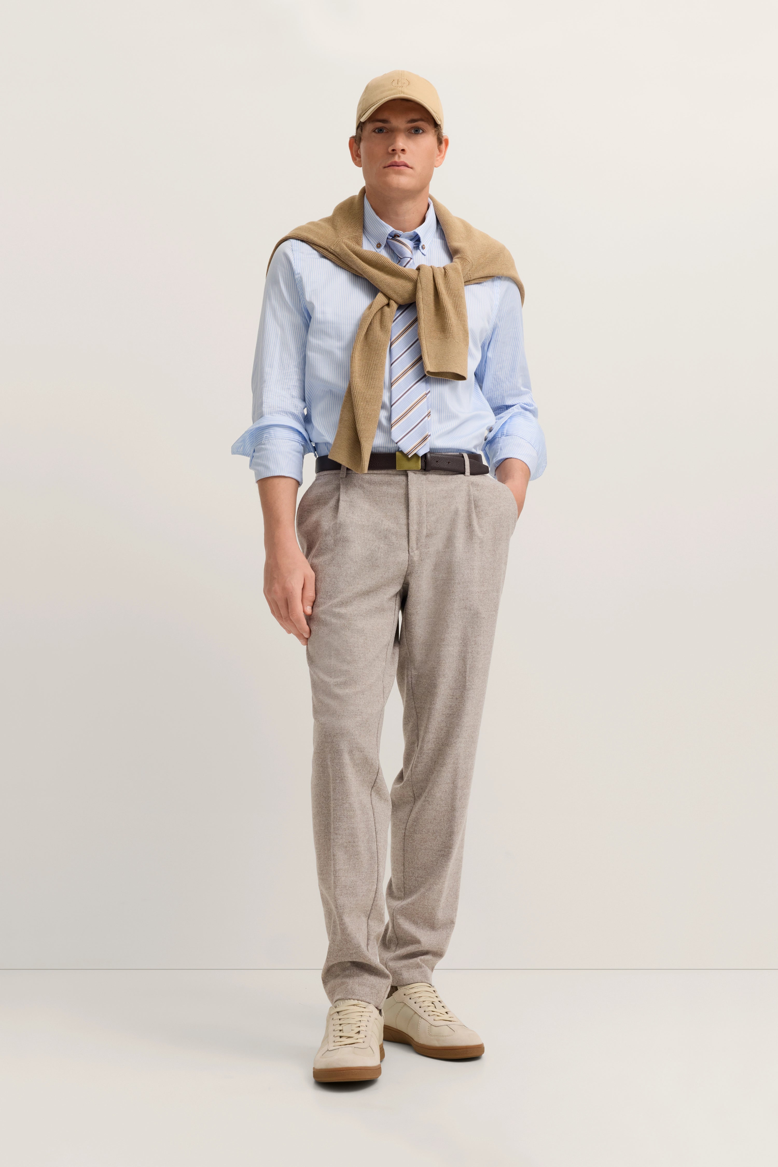 Model in hellblauem Hemd, beige Strickjacke, grauer Hose, Krawatte, Basecap. Lässig mit Händen in Taschen.